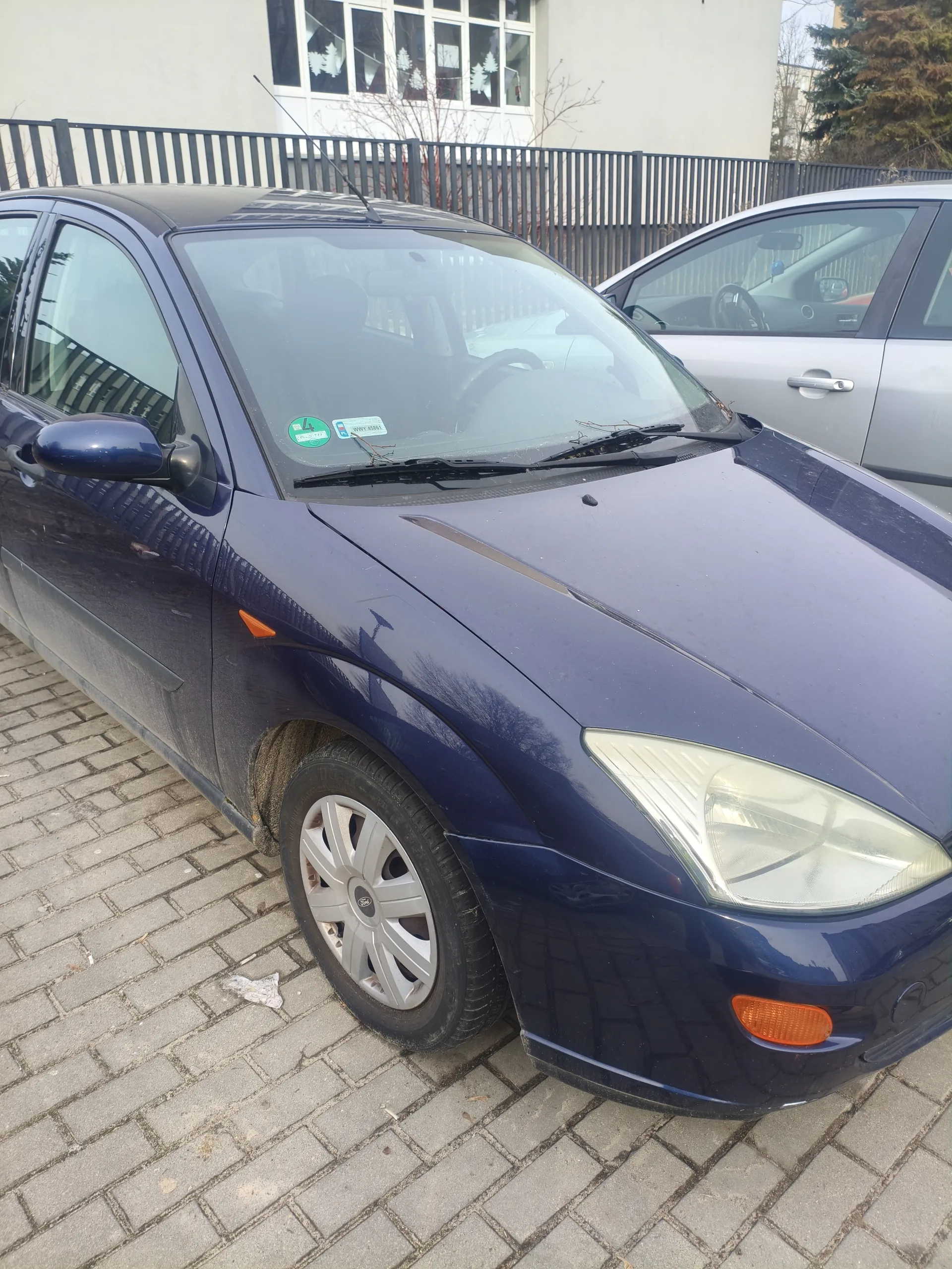 Ciemnoniebieski samochód marki Ford zaparkowany na brukowanej powierzchni. Widoczne są przód pojazdu z reflektorem, pomarańczowym kierunkowskazem i felgą z logo Ford. Na przedniej szybie znajdują się dwa naklejki: zielona z cyfrą 4 oraz niebieska. Obok stoi srebrny samochód. W tle budynek z oknami zdobionymi białymi wzorami, czarny metalowy płot oraz drzewa. Na ziemi leży biały przedmiot.