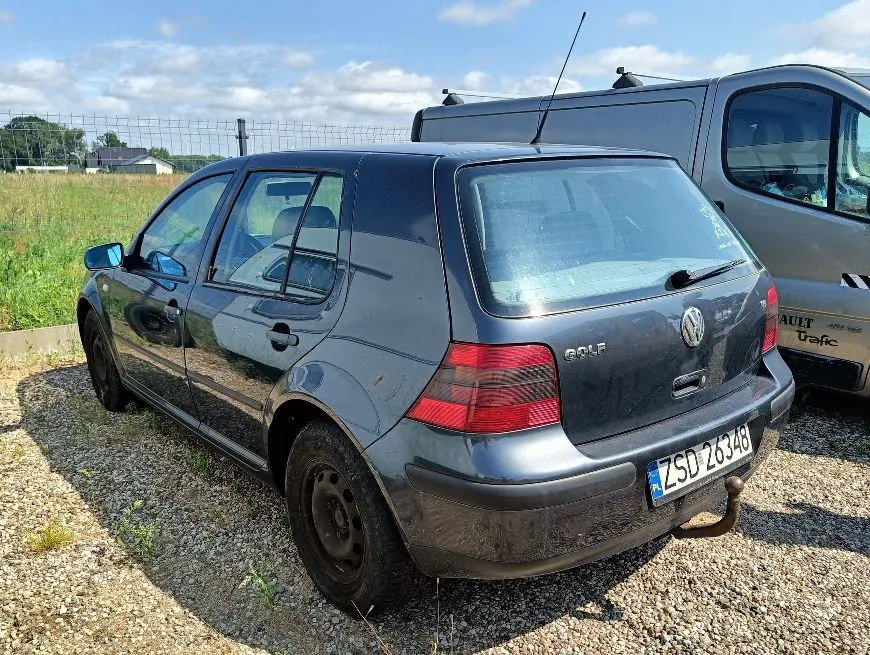 Ciemnoszary samochód osobowy marki Volkswagen Golf zaparkowany tyłem na żwirowym placu. Widoczna jest tylna część pojazdu z tablicą rejestracyjną ZSD 26348, logo Volkswagena oraz napisem GOLF. Obok samochodu stoi biały furgon marki Renault Trafic. W tle znajduje się zielony teren z niską roślinnością i ogrodzenie z siatki pod jasnym, lekko zachmurzonym niebem.