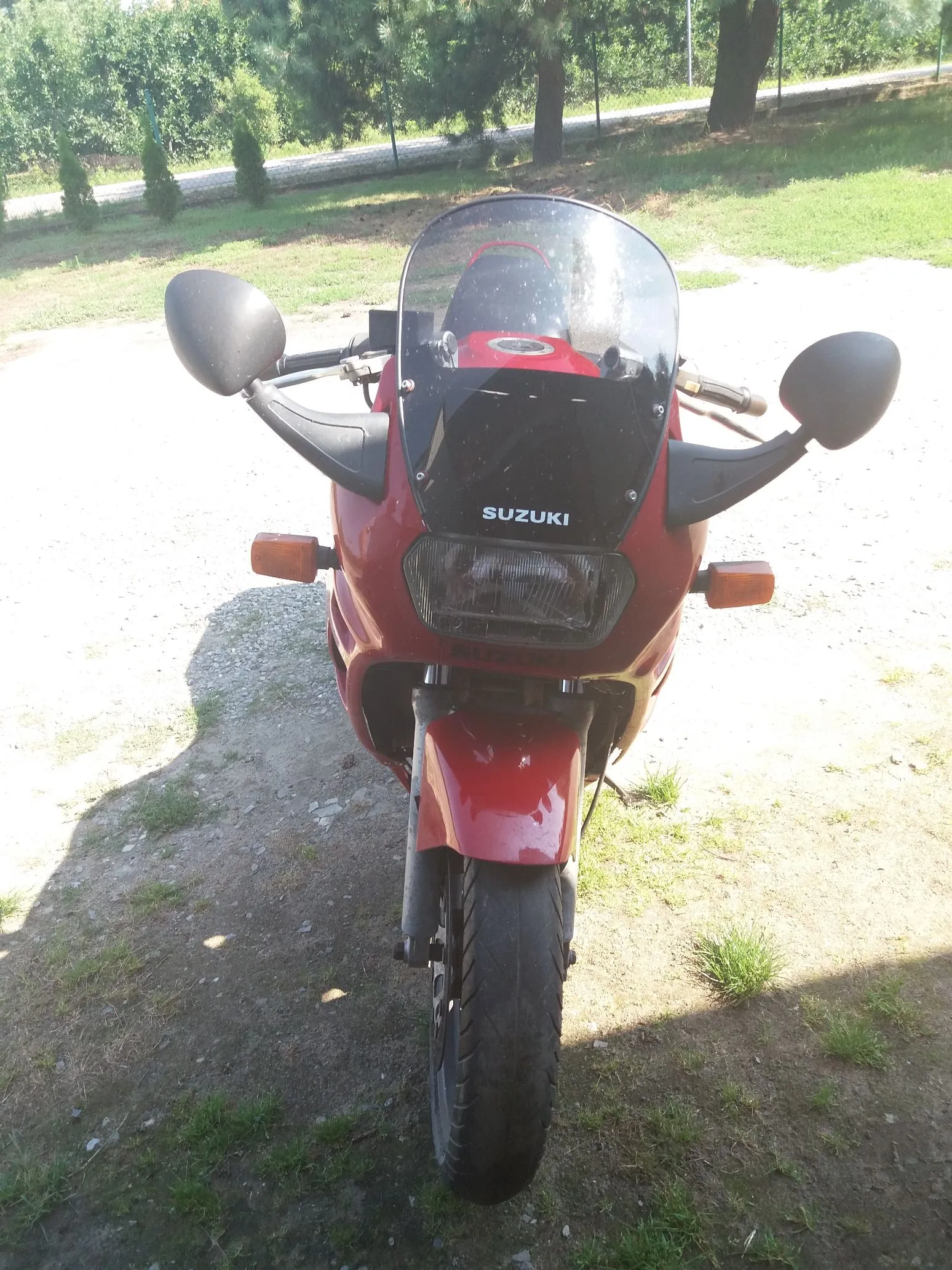 Widok z przodu na czerwony motocykl marki Suzuki zaparkowany na piaszczysto-trawiastej powierzchni. Motocykl posiada czarną szybę z białym napisem SUZUKI, dwa czarne lusterka boczne oraz dwa pomarańczowe kierunkowskazy. W tle widoczne są zielone drzewa i krzewy pod jasnym niebem.