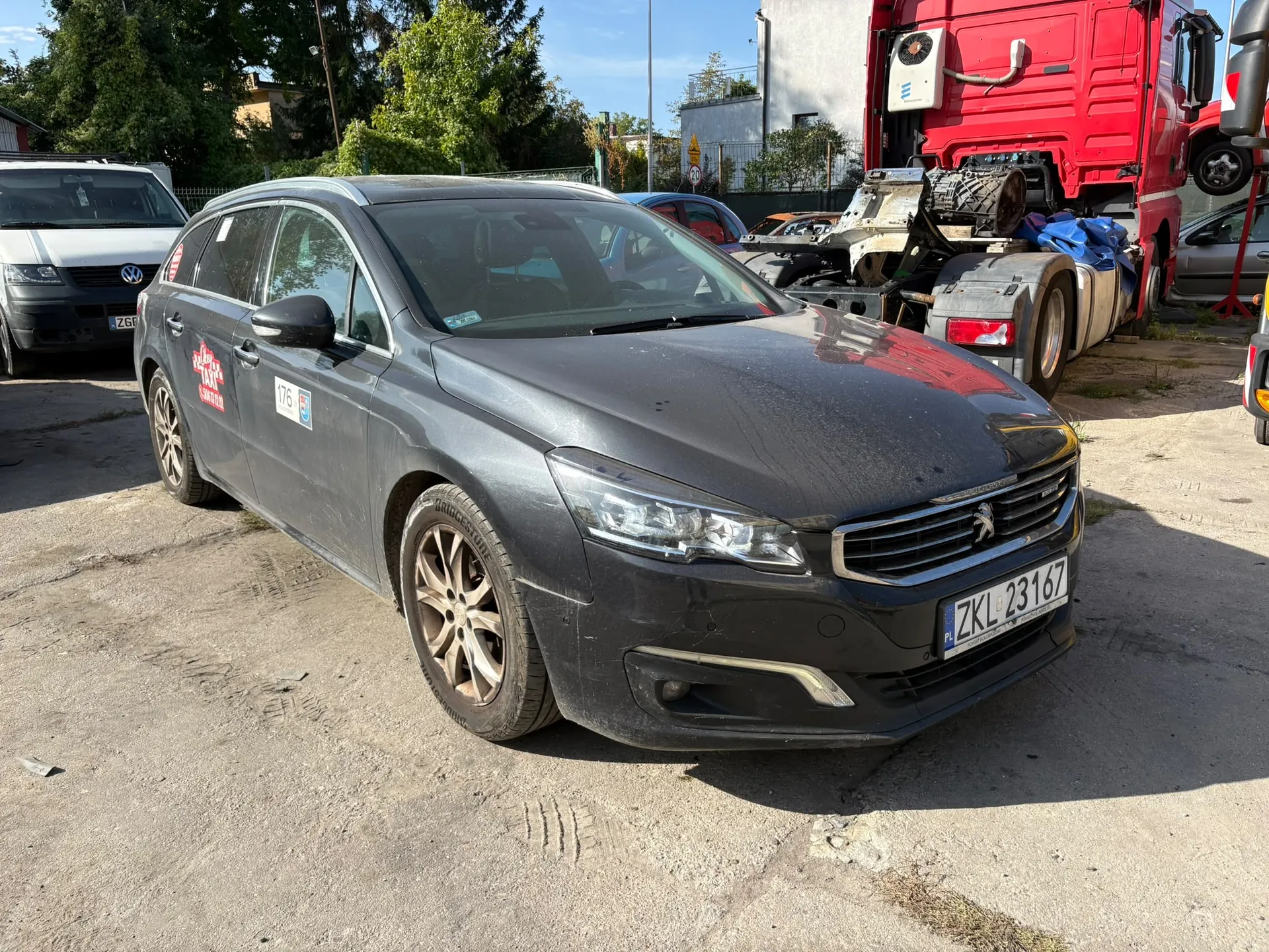 Srebrno-szary samochód marki Peugeot w nadwoziu kombi, zaparkowany na betonowym placu. Auto jest zakurzone i posiada naklejkę z napisem 'rally' na przednich drzwiach oraz numer startowy '176'. W tle widoczny jest czerwony ciągnik siodłowy oraz biały samochód osobowy. W tle znajdują się również drzewa i budynki.