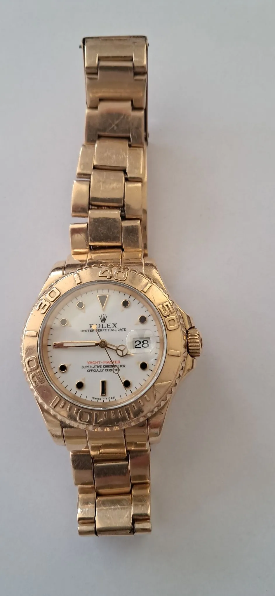 Złoty zegarek marki Rolex model Yacht-Master z metalowym paskiem. Zegarek ma białą tarczę z czarnymi indeksami godzinowymi i wskazówkami. Na tarczy widnieje napis 'ROLEX OYSTER PERPETUAL DATE' oraz 'YACHT-MASTER SUPERLATIVE CHRONOMETER OFFICIALLY CERTIFIED'. Przy godzinie trzeciej znajduje się okienko z datą '28' oraz soczewka powiększająca. Bezel jest złoty z grawerowanymi cyframi 20, 30, 40, 50.