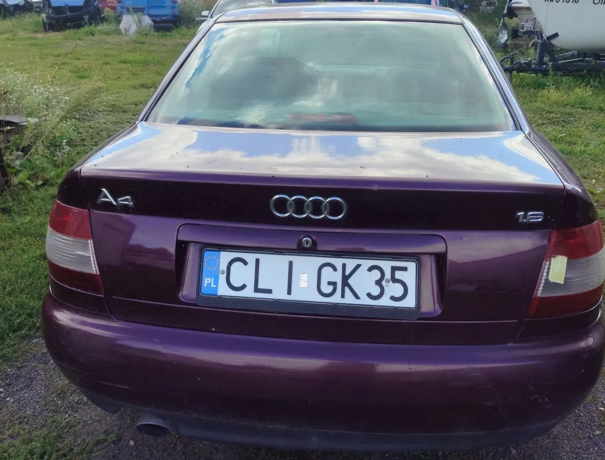 Tylny widok fioletowego samochodu marki Audi A4 1.8. Na klapie bagażnika widoczne jest logo Audi oraz oznaczenia modelu 'A4' i silnika '1.8'. Samochód posiada polską tablicę rejestracyjną o numerze CLI GK35. Pojazd stoi na trawiastym terenie.