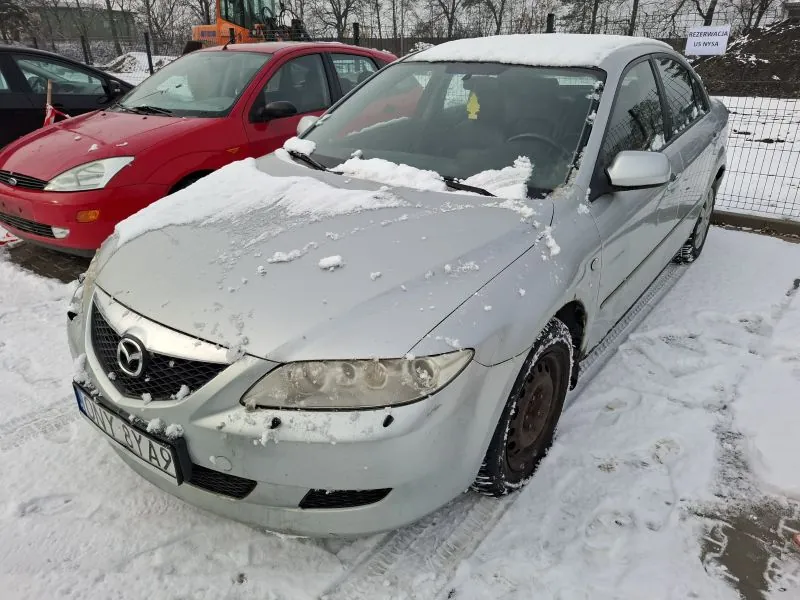 Srebrny samochód marki Mazda z tablicą rejestracyjną WY 6YA9, pokryty śniegiem, zaparkowany na śnieżnej powierzchni. W tle czerwony i czarny samochód, ogrodzenie, drzewa oraz biały znak z napisem 'REZERWACJA 100 M'. Na przedniej szybie srebrnego samochodu widoczny żółty znaczek. Śnieg pokrywa dach i maskę pojazdu.