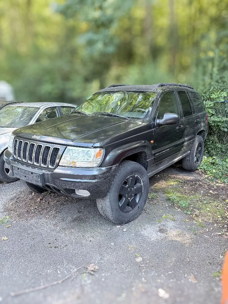 Czarny samochód terenowy marki Jeep Grand Cherokee zaparkowany na szutrowej drodze obok innego, białego samochodu. Pojazd jest zabrudzony błotem, szczególnie w dolnych partiach nadwozia i na kołach. W tle widoczne są zielone drzewa i krzewy.