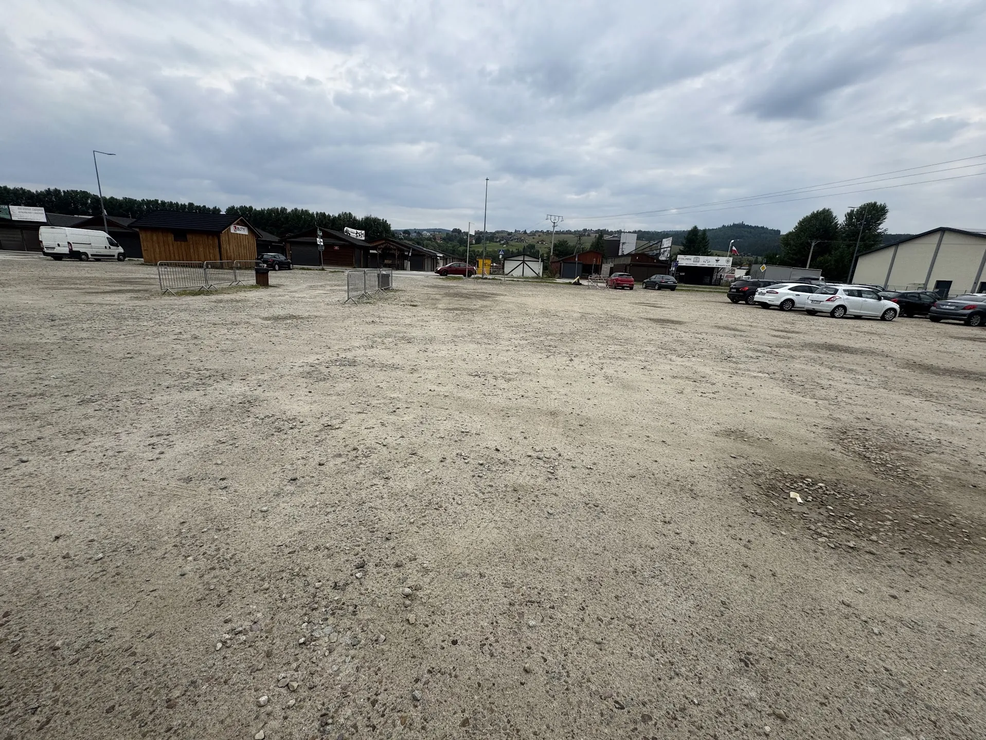 Szeroki widok na nieutwardzony, żwirowy parking pod pochmurnym niebem. W tle znajdują się niskie budynki gospodarcze, magazyny oraz kilka zaparkowanych samochodów osobowych i biały samochód dostawczy. Po lewej stronie widoczne są metalowe barierki odgradzające część terenu. W oddali widać linię drzew i wzgórza.