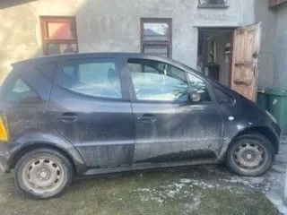 Ciemnoszary, zabrudzony samochód osobowy typu hatchback zaparkowany na trawie przed budynkiem. Samochód ma brudne felgi i widoczne ślady błota na karoserii. W tle budynek z betonową elewacją, dwoma oknami z czerwonymi ramami i otwartymi drewnianymi drzwiami. Po prawej stronie drzwi znajduje się zielony pojemnik na śmieci. Na ziemi widoczne są plamy śniegu lub soli.