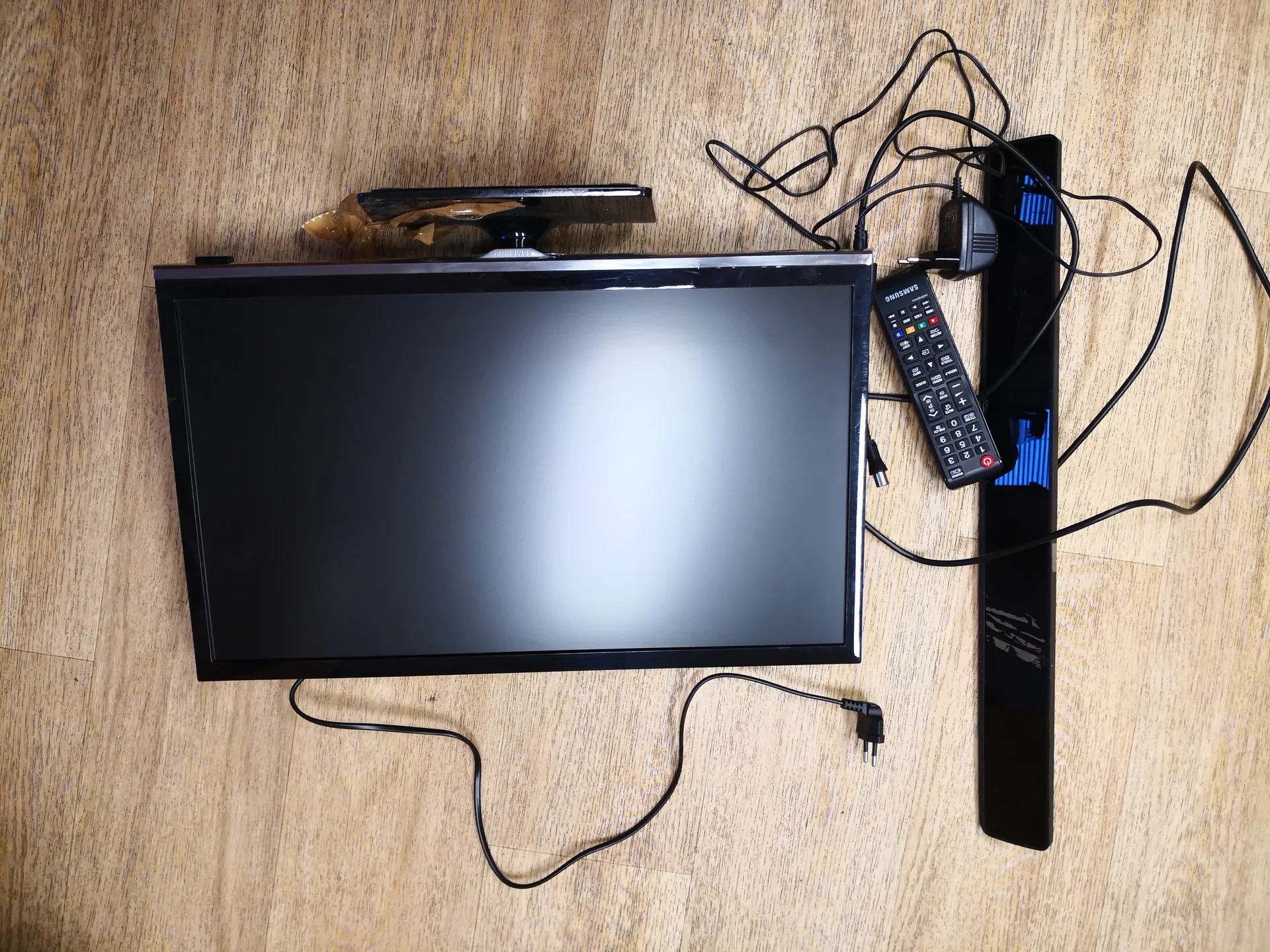 Czarny telewizor płaskoekranowy leżący na podłodze z drewnianym wzorem. Obok telewizora znajduje się czarny pilot z widocznym napisem SAMSUNG, kable elektryczne oraz czarny prostokątny przedmiot z niebieskimi elementami. Ekran telewizora jest wyłączony, a na podłodze widoczne są rozłożone przewody.