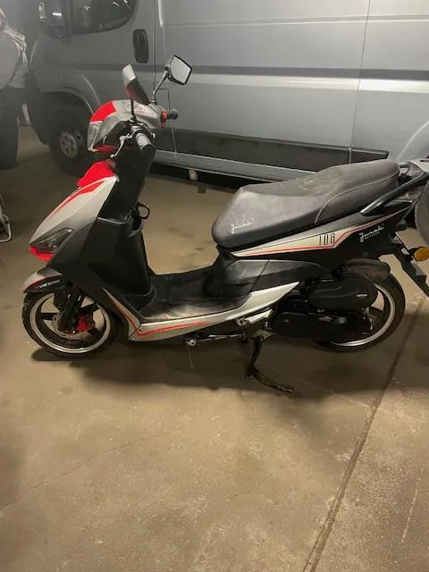 Czarny skuter z czerwonymi i białymi akcentami, stojący na betonowej podłodze. Na boku skutera widoczny napis "100" oraz częściowo widoczny napis "Jog". W tle srebrny furgon. Skuter ma czarne siedzenie, lusterka boczne i czerwone elementy na przednim kole. Podłoga jest szara, a całość znajduje się w pomieszczeniu przypominającym garaż.