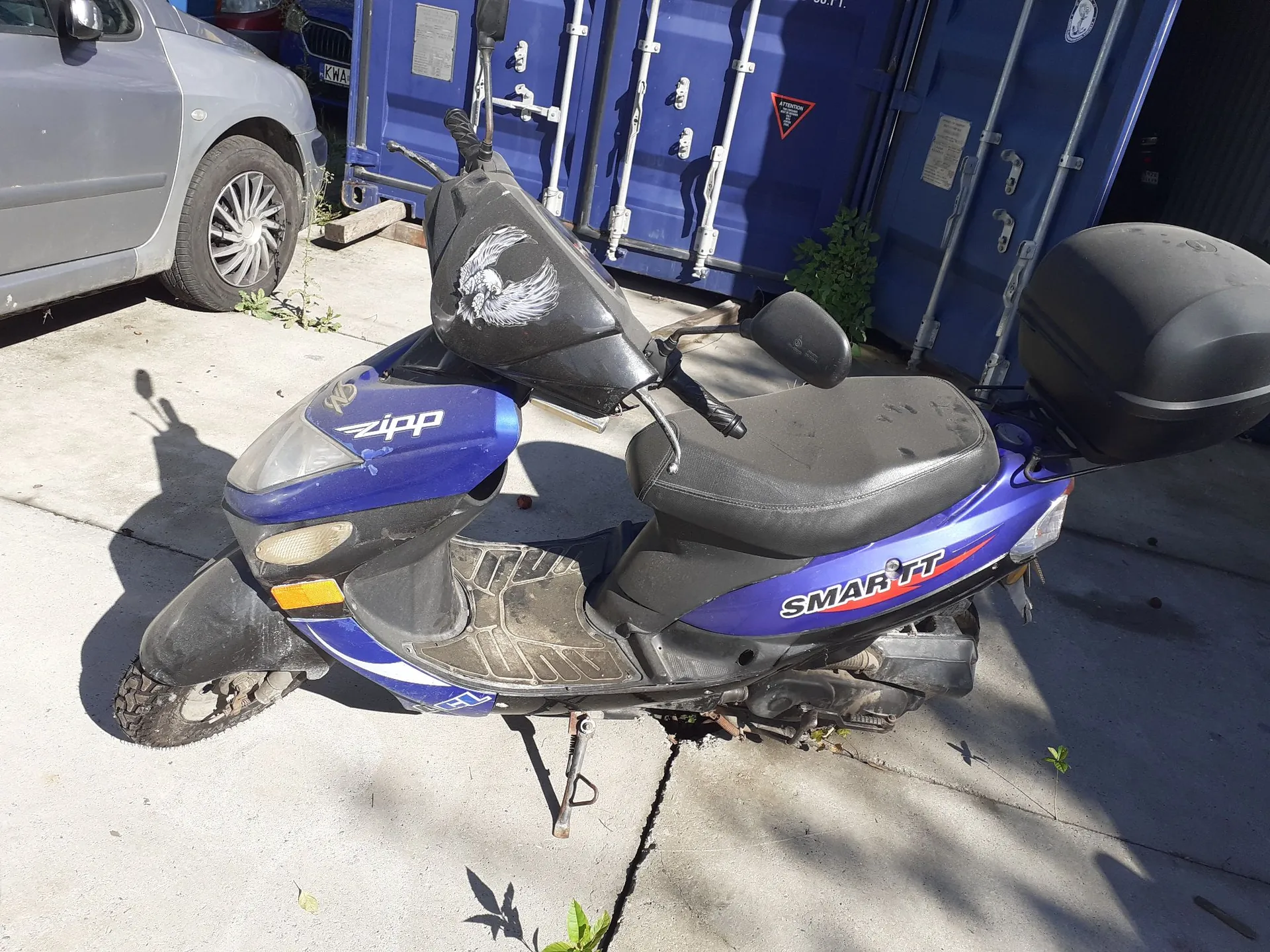 Niebieski skuter marki Zipp z napisem SMART TT na boku, zaparkowany na betonowym podjeździe przed niebieskimi kontenerami transportowymi. Skuter posiada czarną kanapę, czarny schowek z tyłu oraz naklejkę z białym orłem na przedniej osłonie kierownicy.