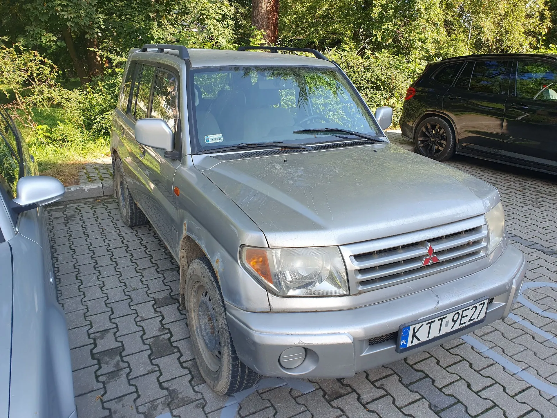 Srebrny samochód terenowy marki Mitsubishi zaparkowany na kostce brukowej. Pojazd ma widoczne ślady zużycia i zabrudzenia na nadwoziu oraz oponach. Na przednim zderzaku znajduje się tablica rejestracyjna o numerze KTT 9E27. W tle widoczne są zielone drzewa oraz fragment czarnego samochodu zaparkowanego obok.