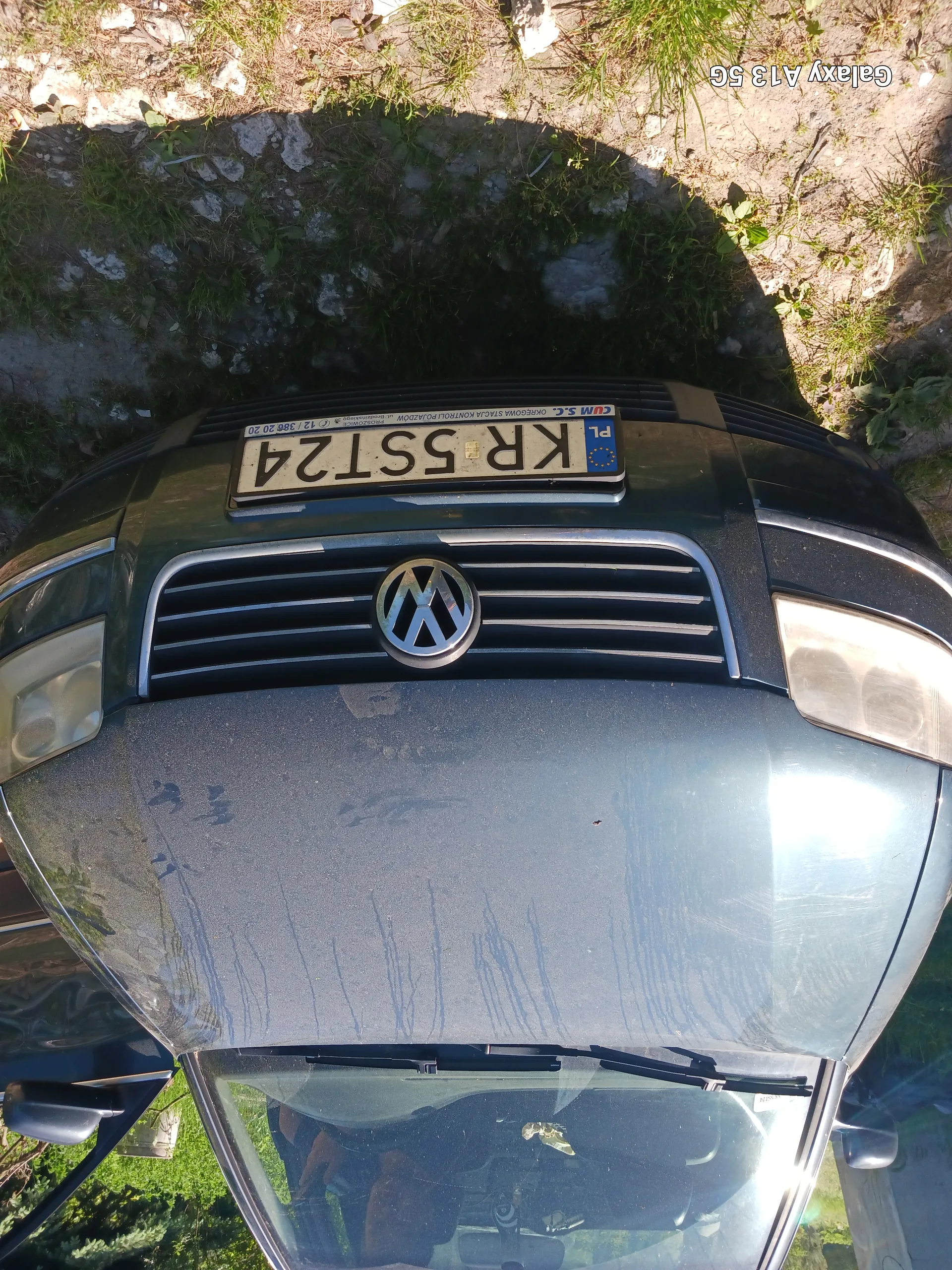 Czarny samochód marki Volkswagen z widocznym logo na atrapie chłodnicy. Tablica rejestracyjna z białym tłem i czarnym tekstem KR 55T24, z niebieskim pasem z napisem PL oraz flagą UE po lewej stronie. Na masce widoczne są zadrapania. Drzwi kierowcy są otwarte. Samochód zaparkowany na podłożu z kamieniami i trawą. W prawym dolnym rogu widoczny napis Galaxy A13 5G.