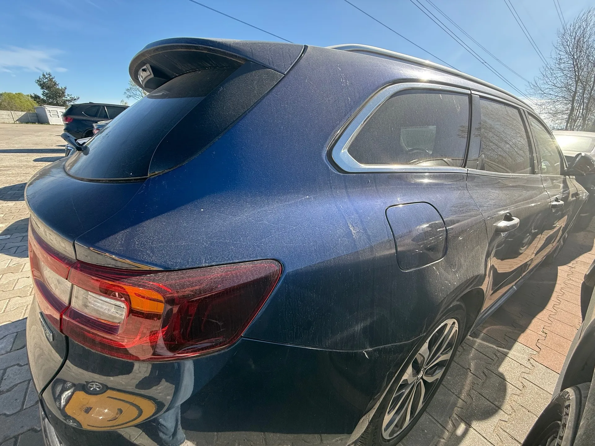 Niebieski samochód typu SUV widziany od tyłu i boku, zaparkowany na brukowanej powierzchni. Widoczne są czerwone światła tylnego oświetlenia, dachowy bagażnik, srebrne felgi oraz żółty naklejony znak z uśmiechniętą buźką na tylnej szybie. W tle znajdują się inne pojazdy, drzewa bez liści, budynek i jasne niebo z kilkoma chmurami. Samochód pokryty jest lekkim kurzem.