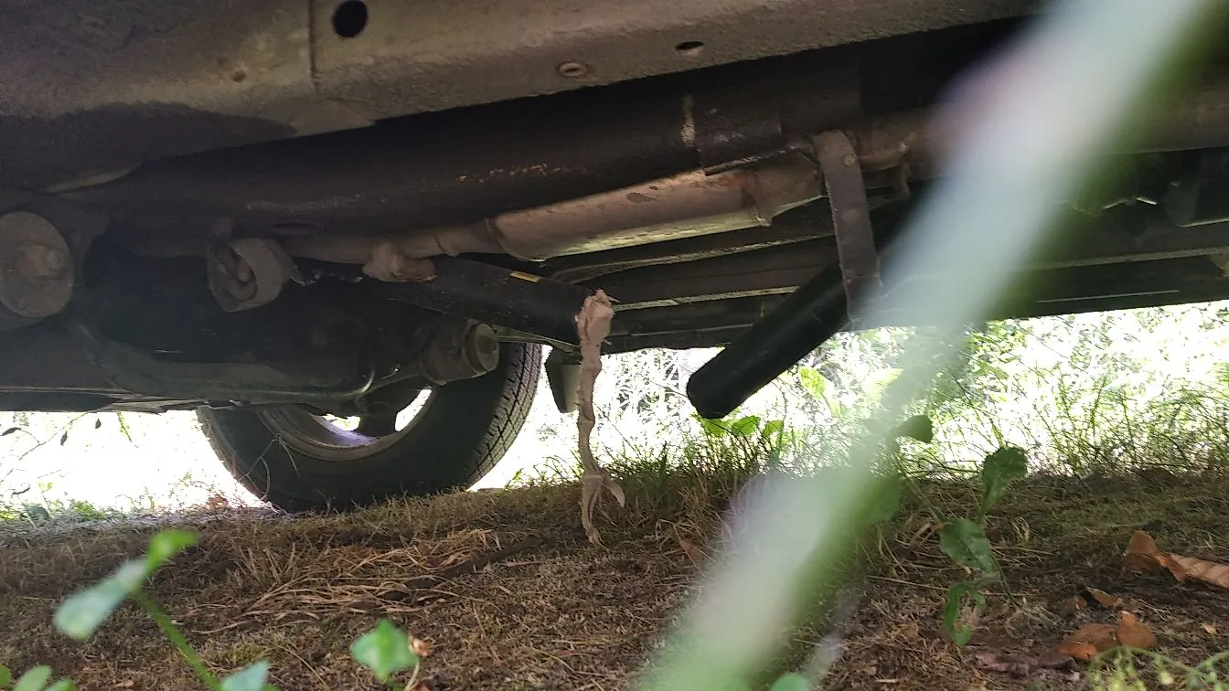 Zdjęcie wykonane z niskiej perspektywy, pokazujące podwozie samochodu. Widoczne są elementy układu wydechowego, w tym rura wydechowa i tłumik, oraz fragment koła z oponą. Z jednej z rur zwisa jasny, poszarpany kawałek materiału lub tkaniny. Tło stanowi ziemia z trawą i roślinnością, a w prawym górnym rogu znajduje się rozmyty element rośliny w pierwszym planie.