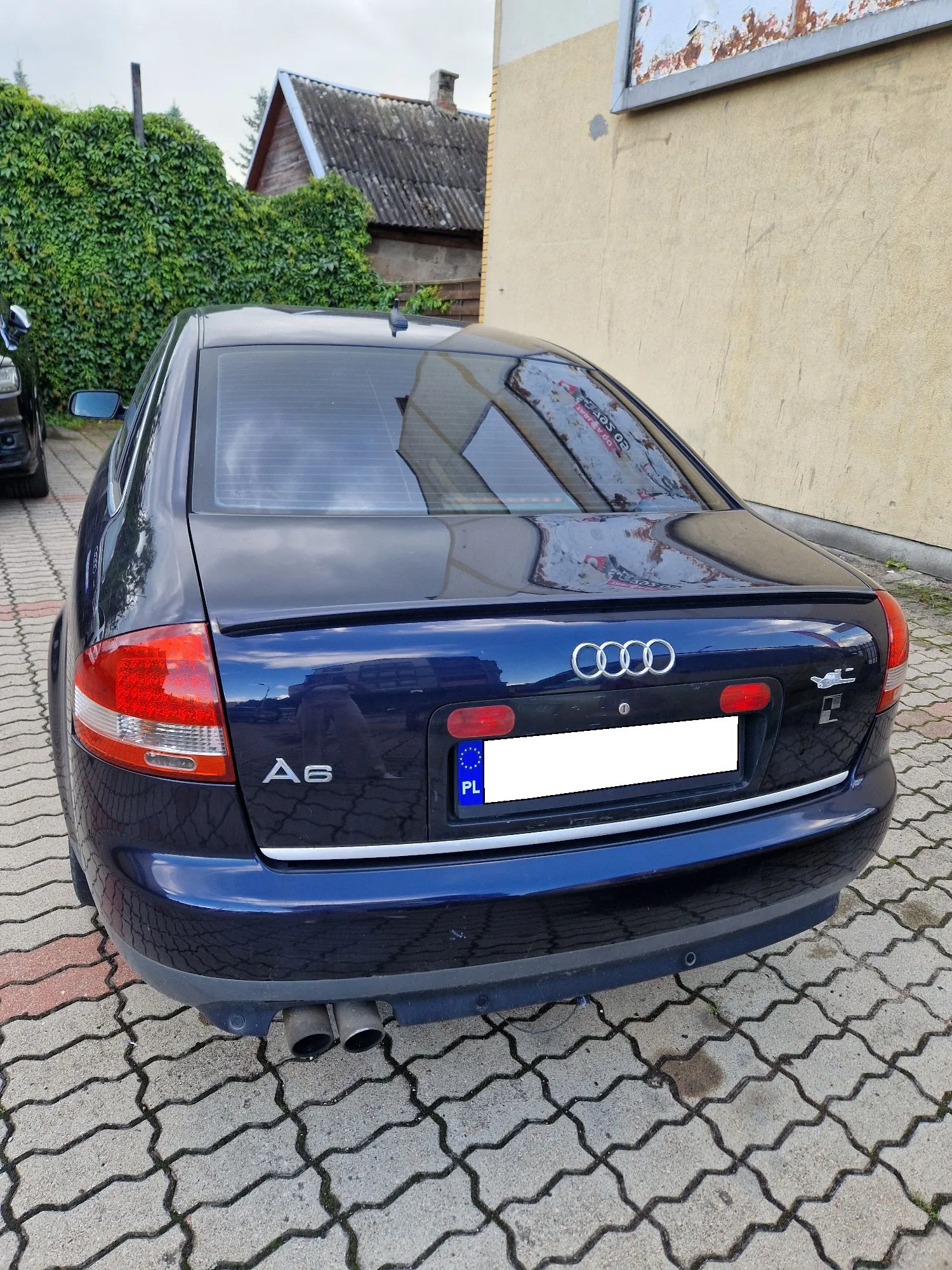 Granatowy samochód Audi A6 zaparkowany tyłem na kostce brukowej. Na klapie bagażnika widoczne jest logo Audi, oznaczenie modelu 'A6' oraz tablica rejestracyjna z oznaczeniem kraju 'PL'. W tle znajduje się żółta ściana budynku oraz zielony żywopłot i fragment dachu starego domu.