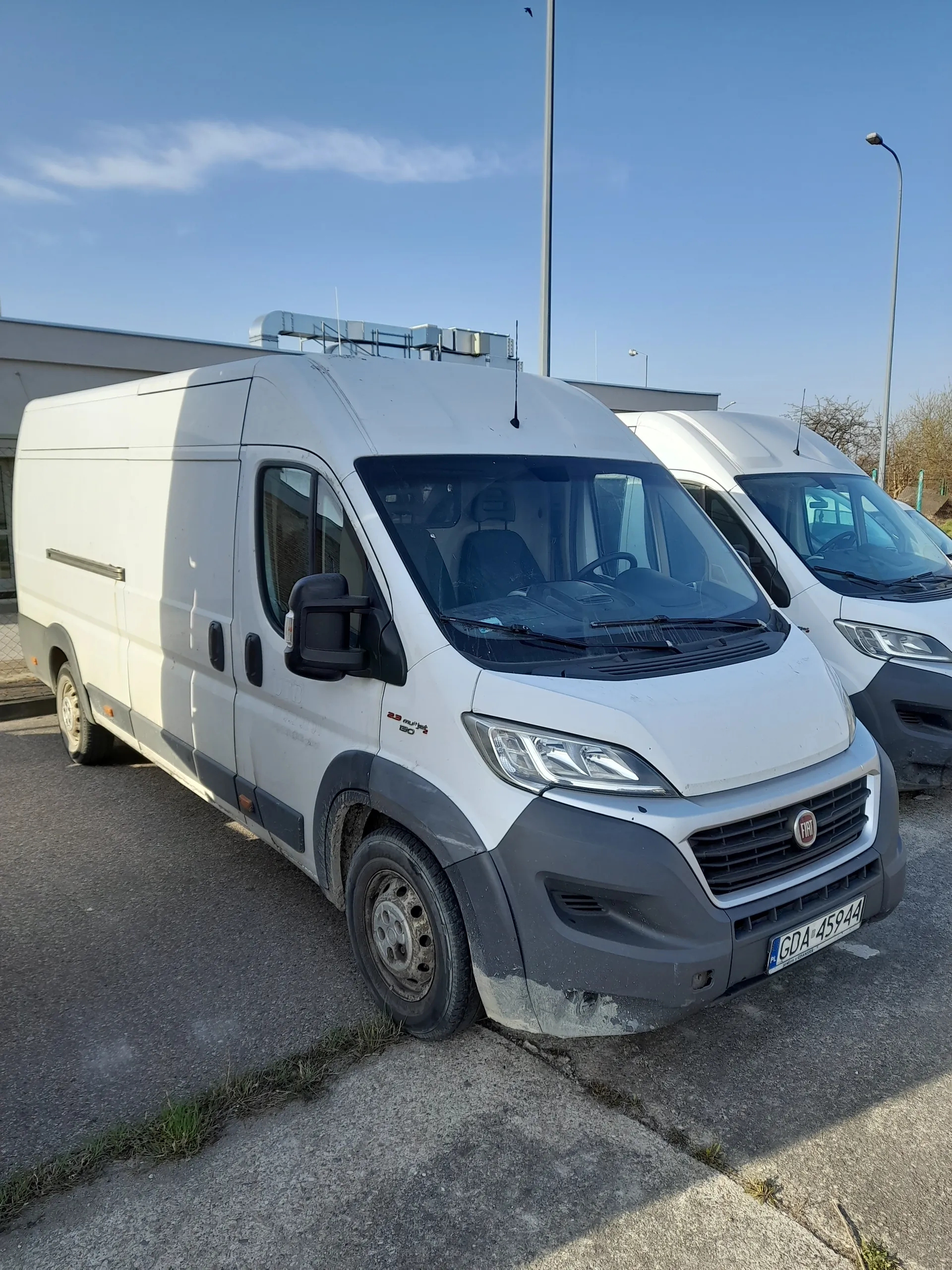Dwa białe furgony zaparkowane na asfalcie. Przedni furgon marki Fiat z tablicą rejestracyjną GDA 4594. W tle budynki, latarnie uliczne oraz niebo z kilkoma chmurami. Fragmenty trawy przy krawężniku na powierzchni asfaltowej.
