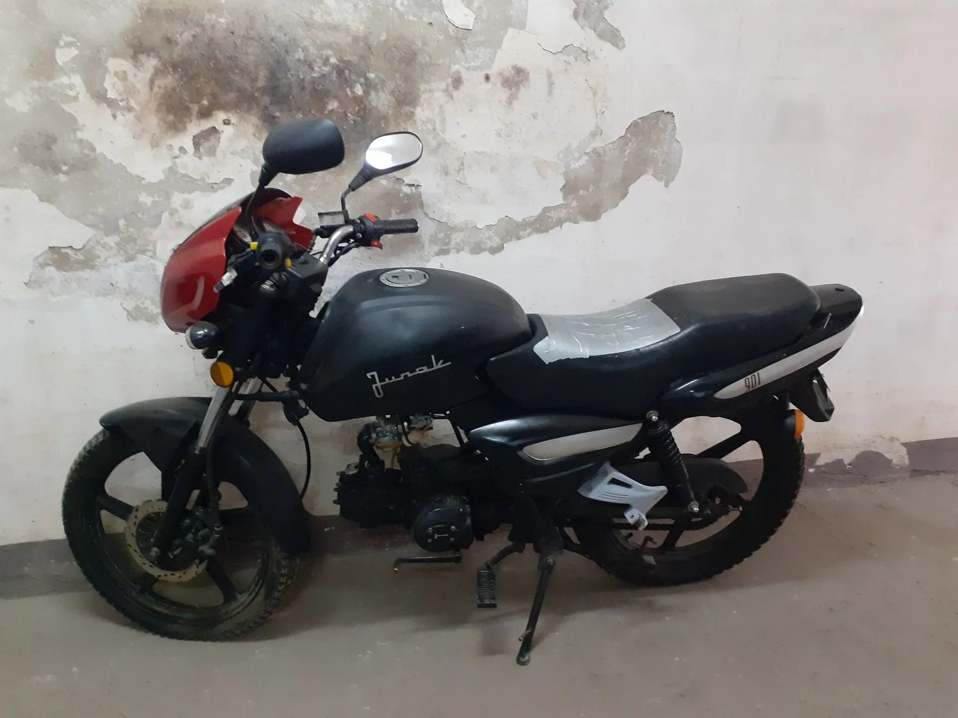 Czarny motocykl marki Junak z czerwonym reflektorem, zaparkowany w pomieszczeniu na tle starej, odrapanej ściany. Motocykl stoi na bocznej stopce, widoczny jest napis 'Junak' na baku paliwa oraz numer '9011' na tylnej części siedzenia.