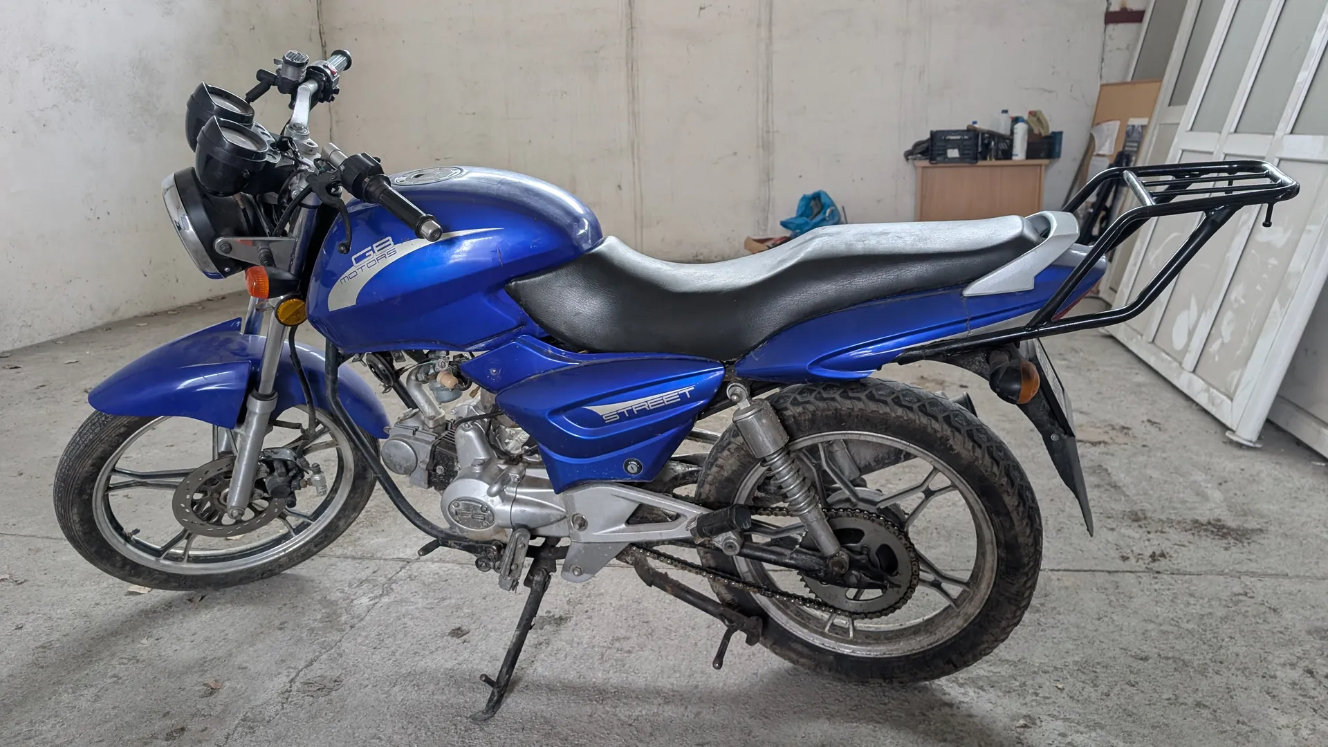 Niebieski motocykl marki CG z napisem 'STREET' na boku, stojący na podwórku lub w garażu. Motocykl ma czarno-srebrne siedzenie, metalowe elementy, reflektory i bagażnik na tył. W tle betonowa ściana, szafka z przedmiotami i białe drzwi. Tekst widoczny na motocyklu: 'STREET' oraz częściowo 'CG' na zbiorniku paliwa.