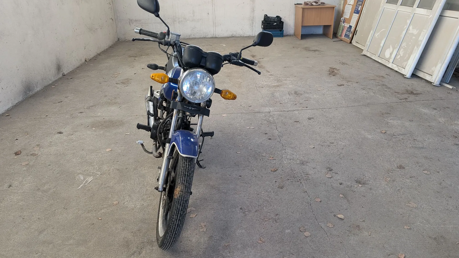 Widok z przodu na niebieski motocykl zaparkowany na betonowym podjeździe. Motocykl posiada okrągły reflektor, dwa pomarańczowe kierunkowskazy oraz czarne lusterka. W tle widoczna jest jasna ściana, biurko oraz kilka białych paneli opartych o ścianę.