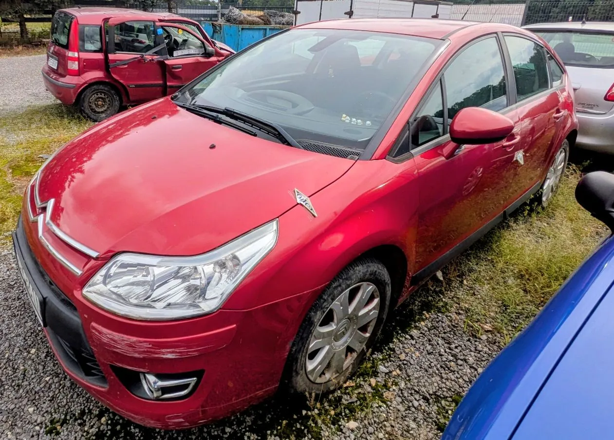Czerwony samochód osobowy marki Citroën zaparkowany na żwirowym podłożu. W tle widoczny jest inny czerwony samochód z otwartymi drzwiami oraz fragment srebrnego auta. Po prawej stronie kadru widoczny jest fragment niebieskiego pojazdu.
