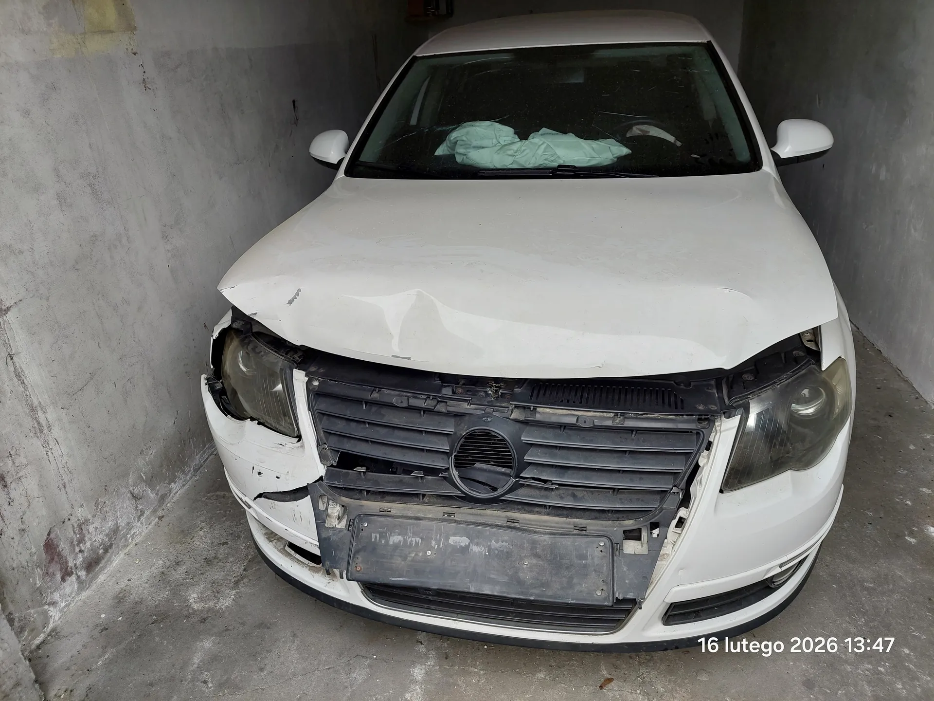 Biały samochód osobowy z uszkodzonym przodem, w tym zdeformowaną maską, pękniętą atrapą chłodnicy i uszkodzonymi reflektorami. Na przedniej szybie leży zielona tkanina. Samochód zaparkowany w betonowym garażu lub podjeździe. W prawym dolnym rogu widoczny tekst: 16 lutego 2026 13:47. Brak tablicy rejestracyjnej.