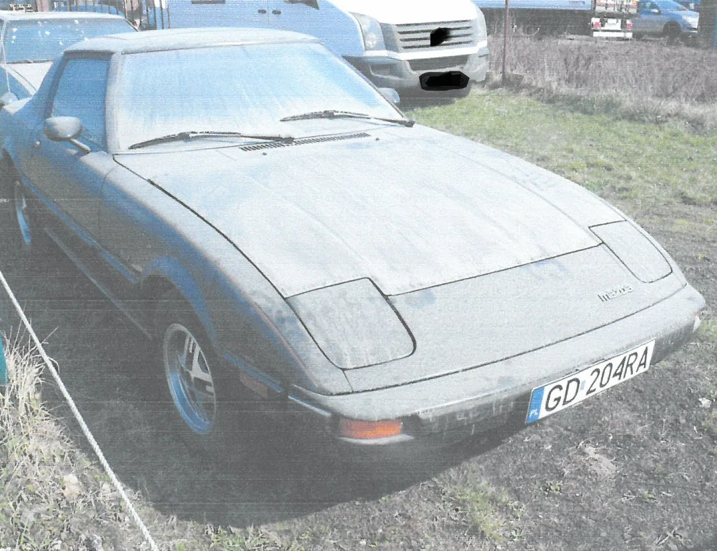 Srebrny samochód sportowy z lat 80. zaparkowany na trawiastym terenie. Pojazd ma chowane reflektory i polską tablicę rejestracyjną o numerze GD 204RA. W tle widoczne są inne samochody.