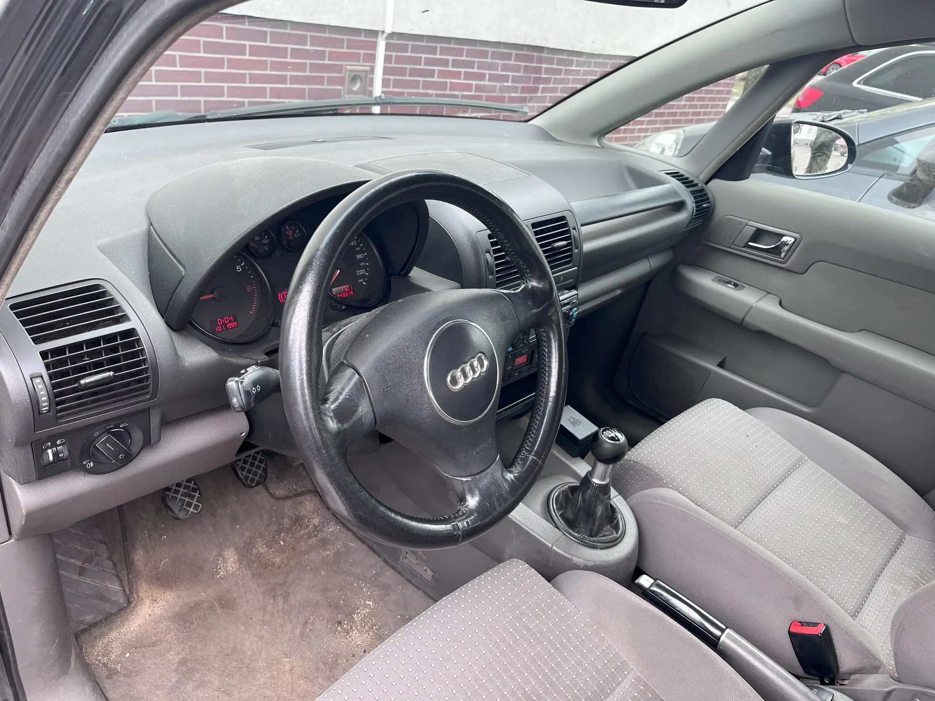 Wnętrze samochodu marki Audi z widocznym kierownicą z logo Audi, szarym deski rozdzielczej, licznikami z czerwonymi cyframi (0:04, 101 1599), manualną skrzynią biegów, szarymi siedzeniami z kropkowanym wzorem oraz widokiem na ceglaną ścianę i inne pojazdy przez przednią szybę.