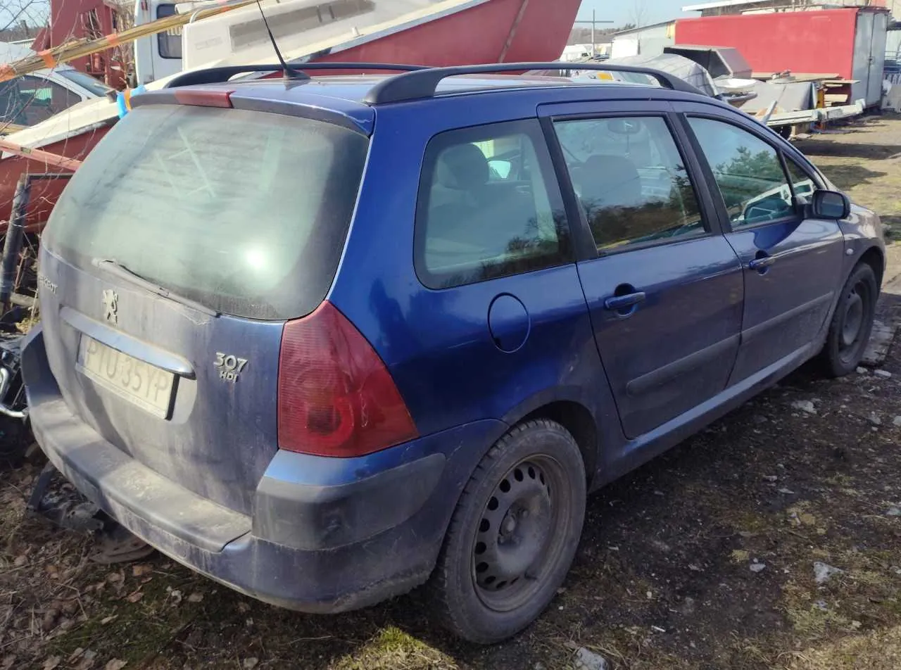 Niebieski samochód osobowy typu kombi marki Peugeot 307 HDI, widok z tyłu i boku. Samochód jest zabrudzony, z widoczną tablicą rejestracyjną PTU 35YP. Na tle znajdują się inne pojazdy, łodzie oraz budynki. Podłoże ziemne z suchą trawą i kamieniami. Słońce oświetla scenę, tworząc cienie.