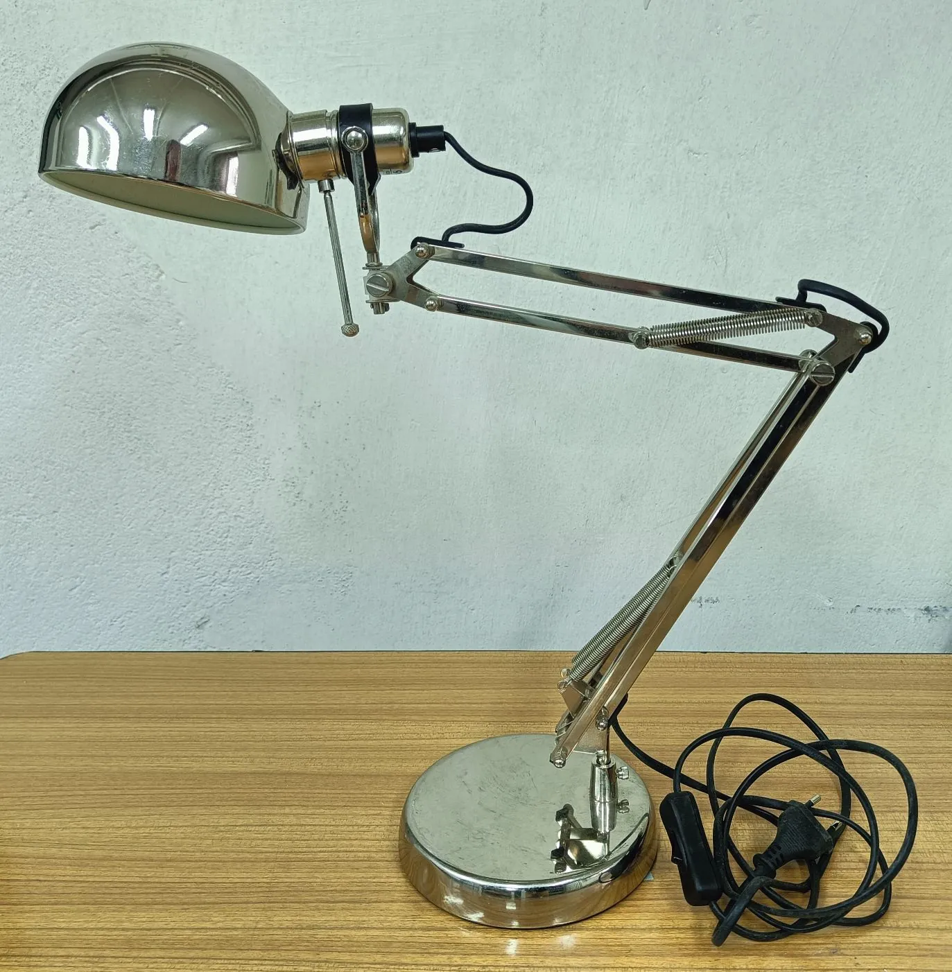 Chromowana lampka biurkowa z regulowanym ramieniem typu pantograf, ustawiona na drewnianym blacie na tle białej ściany. Lampa posiada półkolisty klosz, metalową podstawę w kształcie dysku oraz czarny kabel z wtyczką leżący obok podstawy.