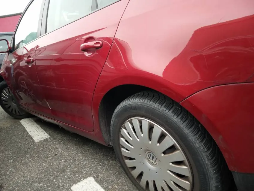 Czerwony samochód Volkswagen widziany z boku, z tylnym kołem z srebrnym felgiem z logo VW, uchwytem drzwiowym oraz częścią innego samochodu na tle. Asfalt z białymi liniami parkingu.
