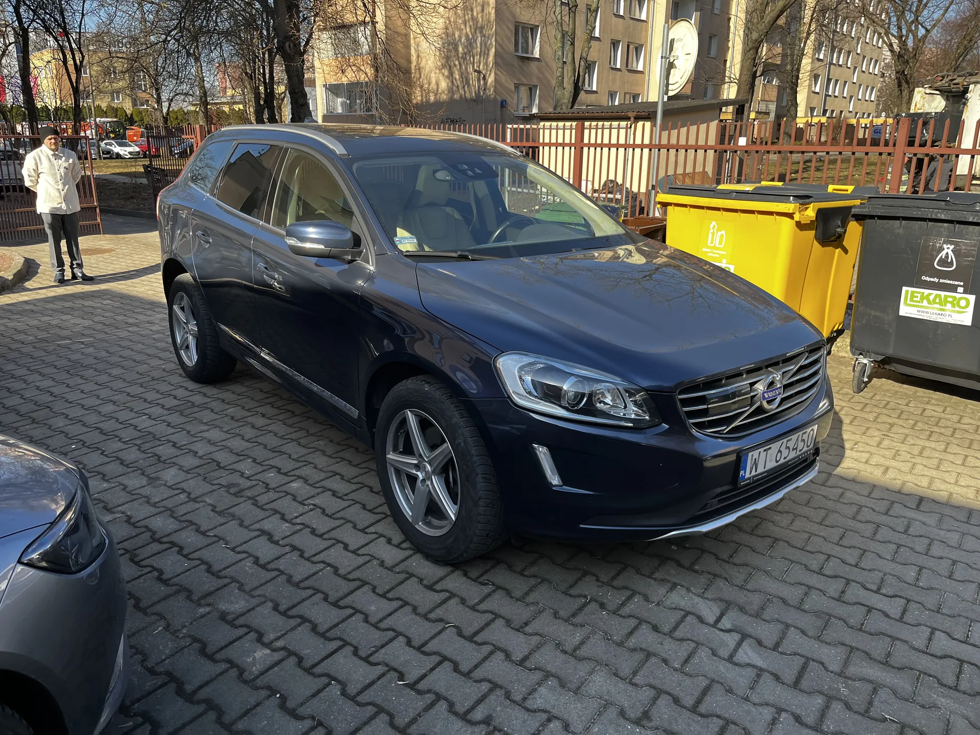 Ciemnoniebieski samochód osobowy marki Volvo z tablicą rejestracyjną WT 65450 zaparkowany na brukowanej powierzchni. Po prawej stronie samochodu znajdują się dwa pojemniki na odpady: żółty z symbolem recyklingu i czarny z napisem LEKARO oraz dodatkowym tekstem w języku polskim. W tle widoczne są budynki mieszkalne, drzewa bez liści, czerwone ogrodzenie oraz osoba stojąca w białej kurtce i ciemnych spodniach. Na lewym dolnym rogu częściowo widoczny jest inny pojazd.