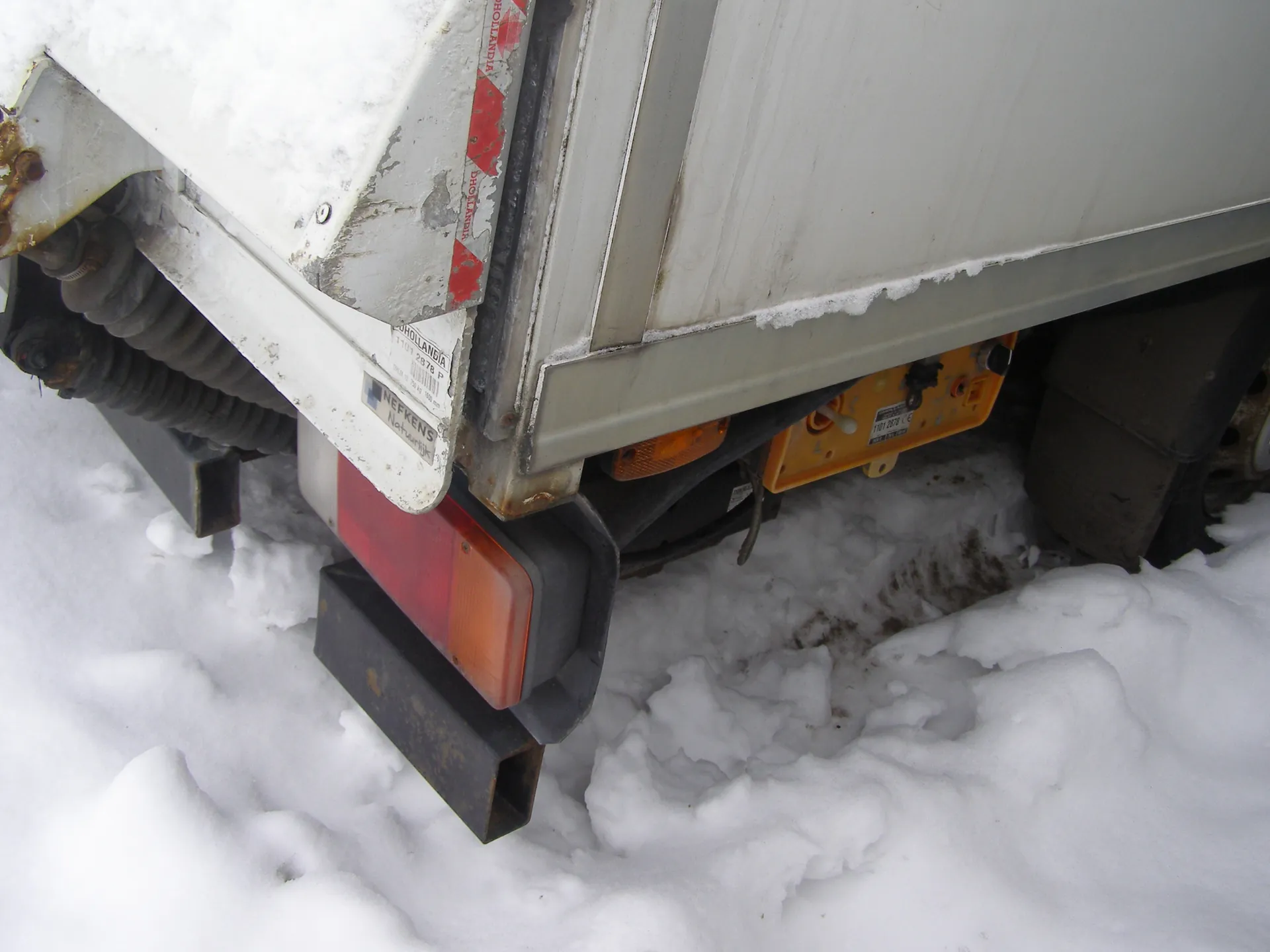 Tył pojazdu ciężarowego z białą przyczepą pokrytą śniegiem. Widoczne czerwone i pomarańczowe światło tylne, rdza na metalowych elementach, naklejki z napisami „NEFKENS” i „HOLLANDIA”. Żółta tabliczka rejestracyjna, śnieg na ziemi z odciskami opon, czarna osłona i sprężyny pod przyczepą.
