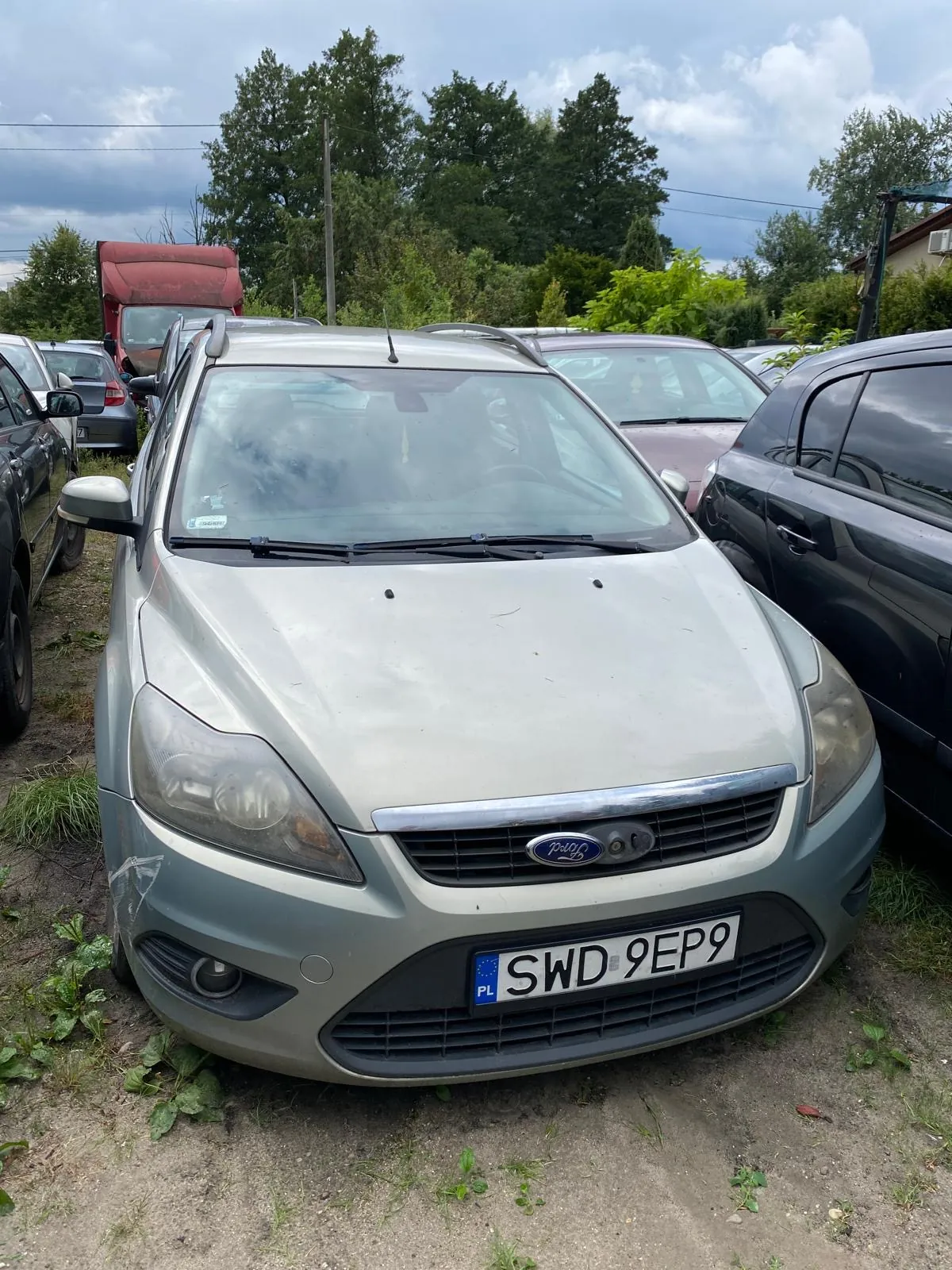 Srebrny samochód marki Ford Focus zaparkowany na piaszczystym placu wśród innych pojazdów. Samochód ma widoczne uszkodzenia karoserii po prawej stronie przedniego zderzaka. Tablica rejestracyjna ma numer SWD 9EP9 i oznaczenie PL.