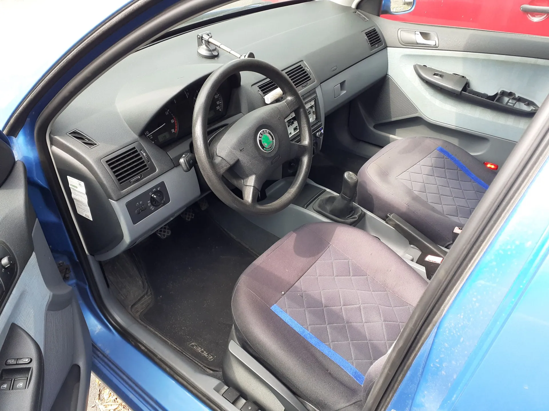 Wnętrze samochodu marki Skoda widziane z perspektywy otwartych drzwi kierowcy. Widoczna jest czarna kierownica z logo marki, deska rozdzielcza z licznikiem i nawiewami, dwa przednie fotele z ciemną tapicerką w romby i niebieskimi wstawkami oraz czarne dywaniki z napisem 'fabia'.