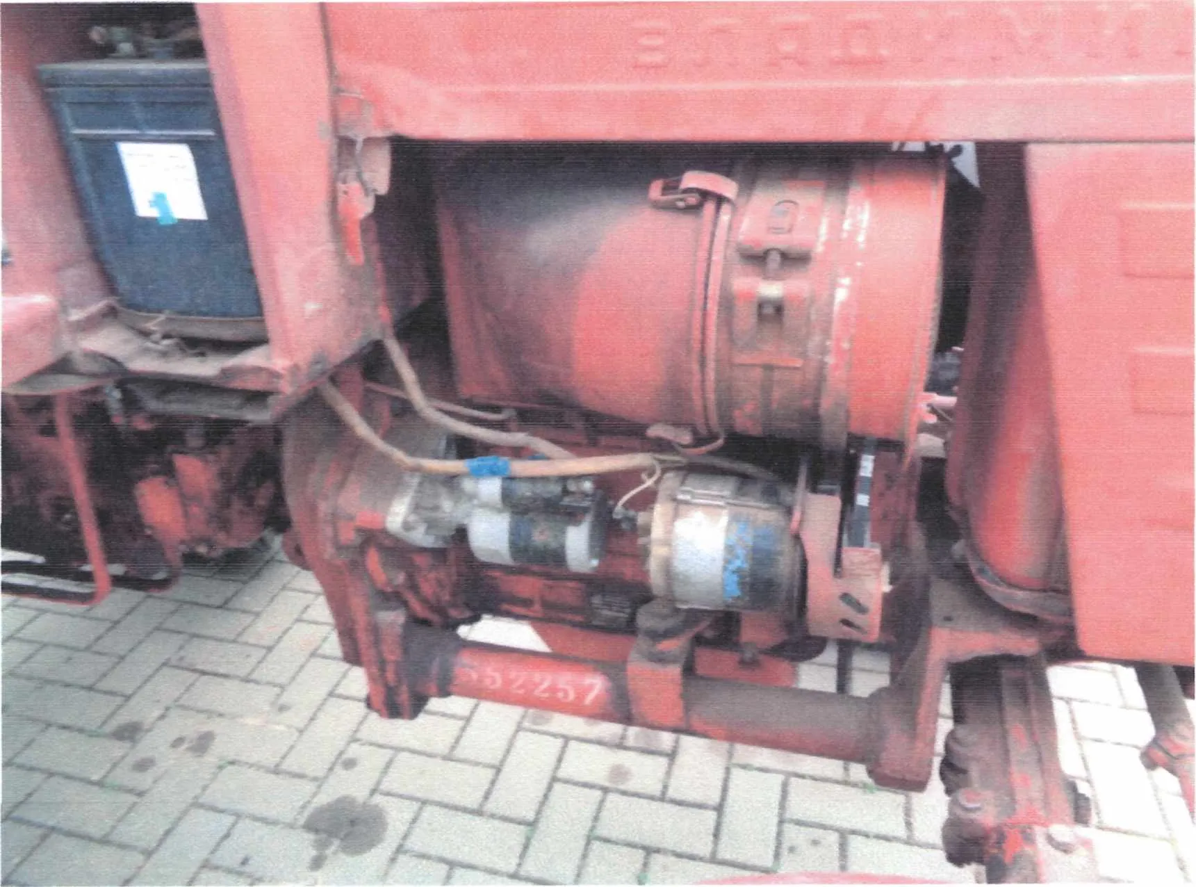 Zdjęcie przedstawia czerwony, przemysłowy obiekt mechaniczny, prawdopodobnie część maszyny lub pojazdu. W centralnej części widoczny jest duży, cylindryczny zbiornik z metalowymi elementami mocującymi i przewodami. Na czerwonej konstrukcji widnieje biały numer "552257". Po lewej stronie znajduje się niebieski pojemnik z białą etykietą i niebieskim znacznikiem. Tło stanowi brukowana powierzchnia z widocznymi plamami. Na górnej części obiektu widoczny jest fragment napisu w języku chińskim.