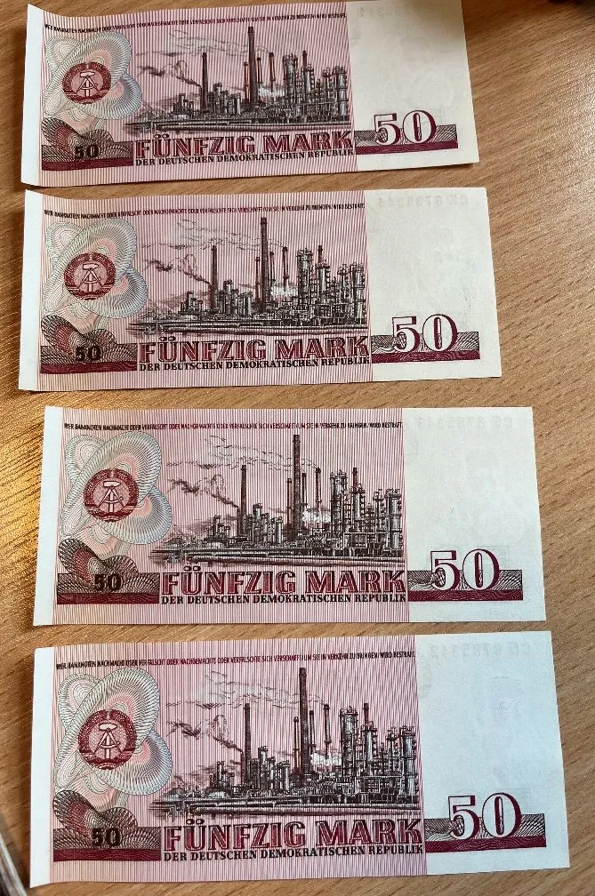 Cztery banknoty o nominale 50 marek z Niemieckiej Republiki Demokratycznej (NRD), ułożone pionowo jeden pod drugim na drewnianym blacie. Banknoty mają różowy kolor i przedstawiają rysunek kompleksu przemysłowego z kominami i fabrykami. Na każdym widnieje napis 'FÜNFZIG MARK' oraz mniejszy tekst 'DER DEUTSCHEN DEMOKRATISCHEN REPUBLIK' oraz liczba '50'.
