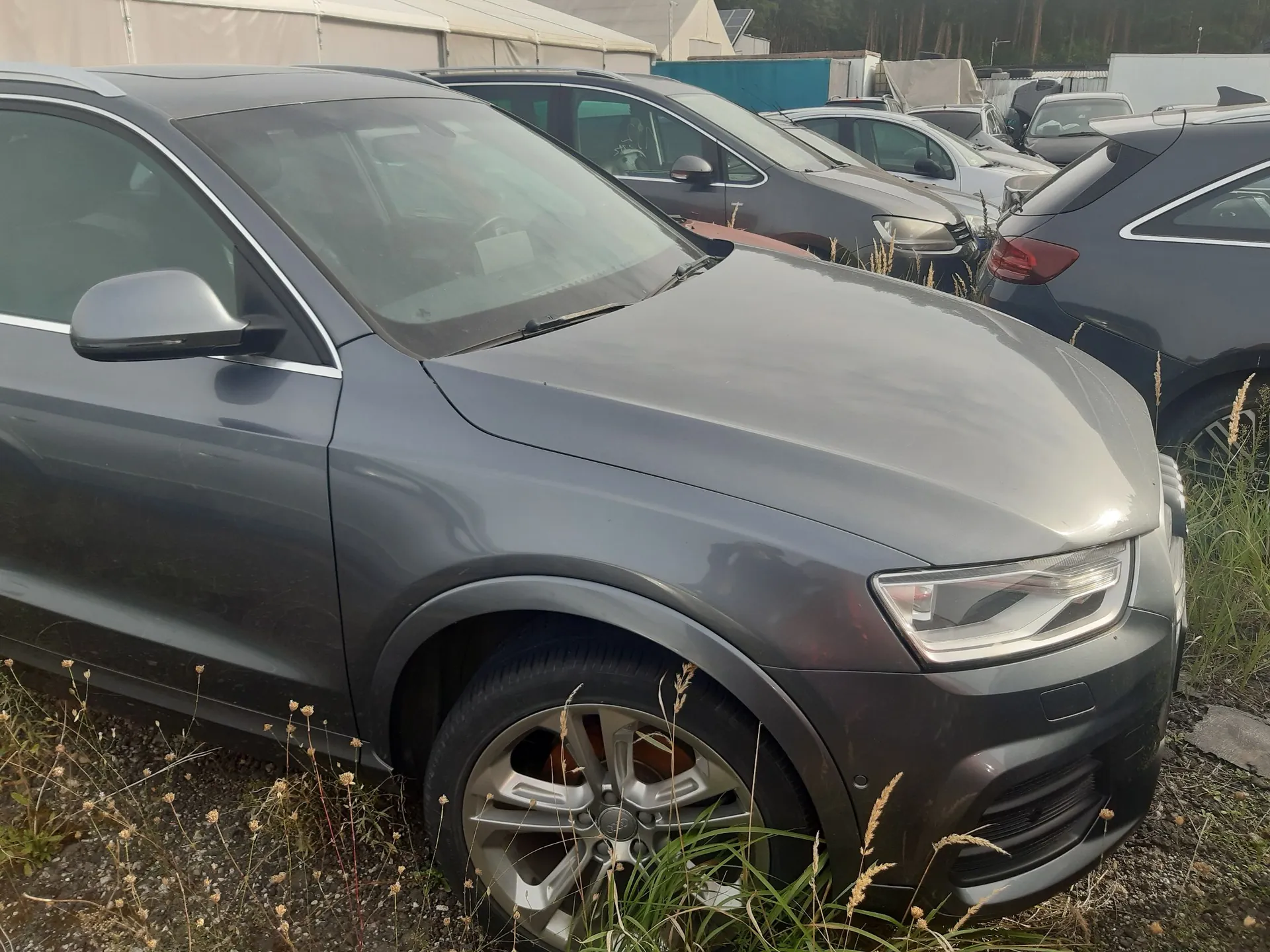 Szary samochód osobowy typu SUV zaparkowany na trawiastym terenie. Widoczny jest przód i bok pojazdu z bliska. W tle znajdują się inne zaparkowane samochody oraz białe namioty magazynowe.