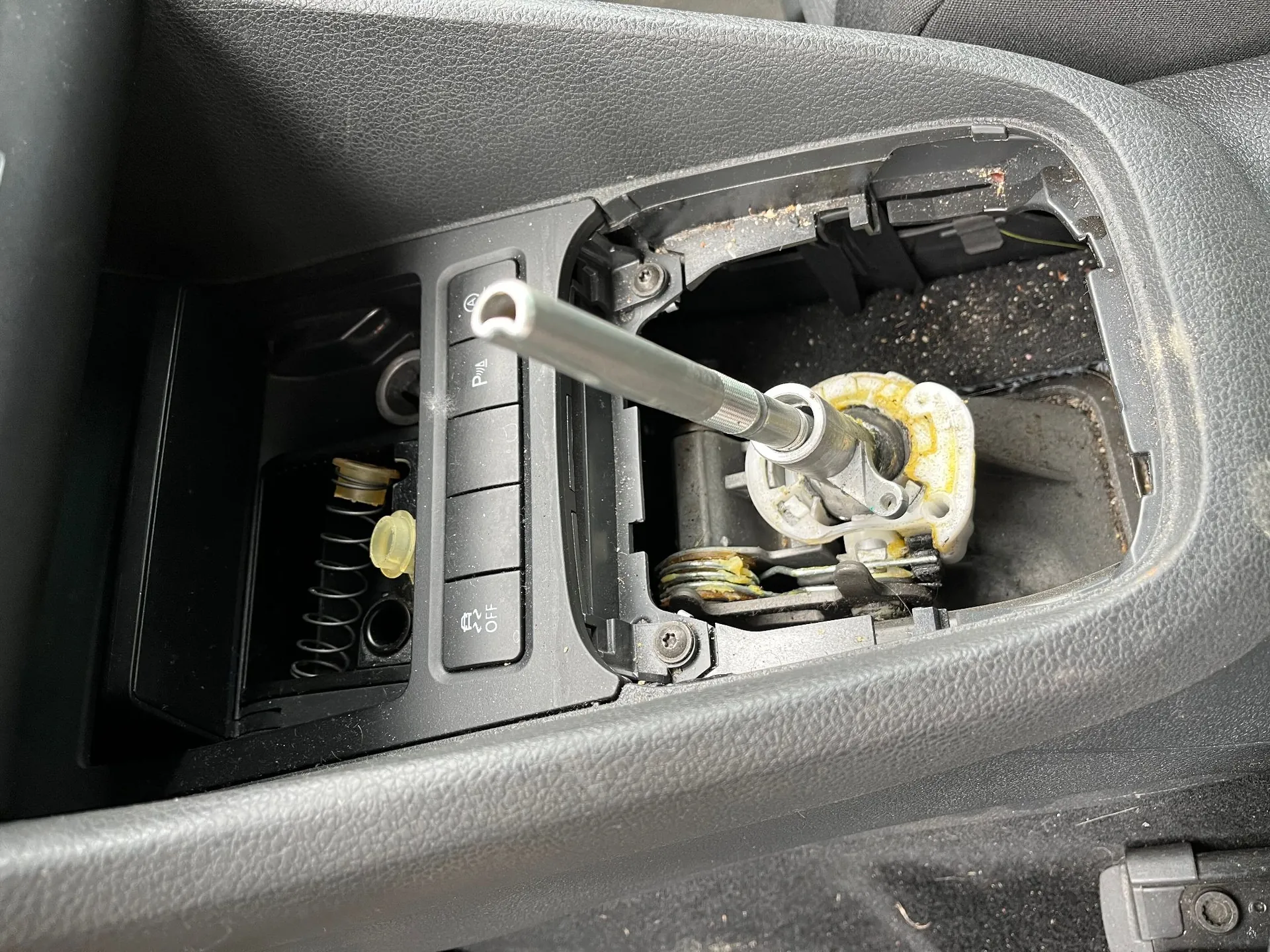 Zbliżenie na wnętrze konsoli środkowej samochodu z wyjętym panelem wykończeniowym. Widoczny jest mechanizm zmiany biegów z metalowym prętem oraz sprężyną w sąsiednim schowku. Po lewej stronie znajdują się przyciski sterujące, w tym przycisk z napisem 'P' oraz przycisk z ikoną i napisem 'OFF'.