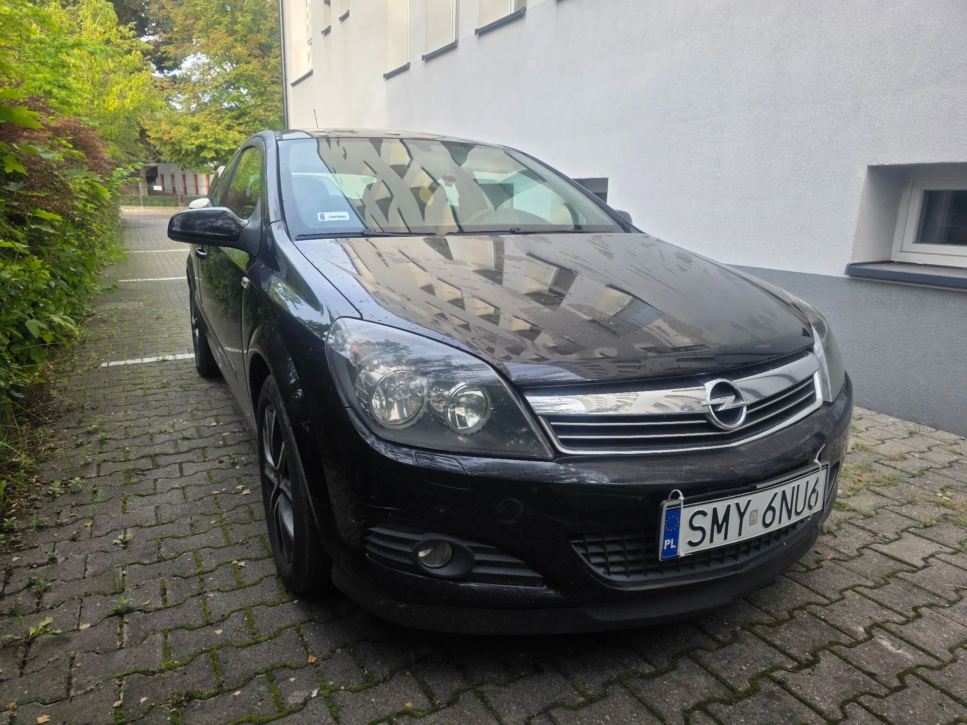 Czarny samochód marki Opel zaparkowany na kostce brukowej obok białego budynku. Widoczna jest przednia część pojazdu z tablicą rejestracyjną o numerze SMY 6NU6. W tle znajdują się zielone krzewy i drzewa.