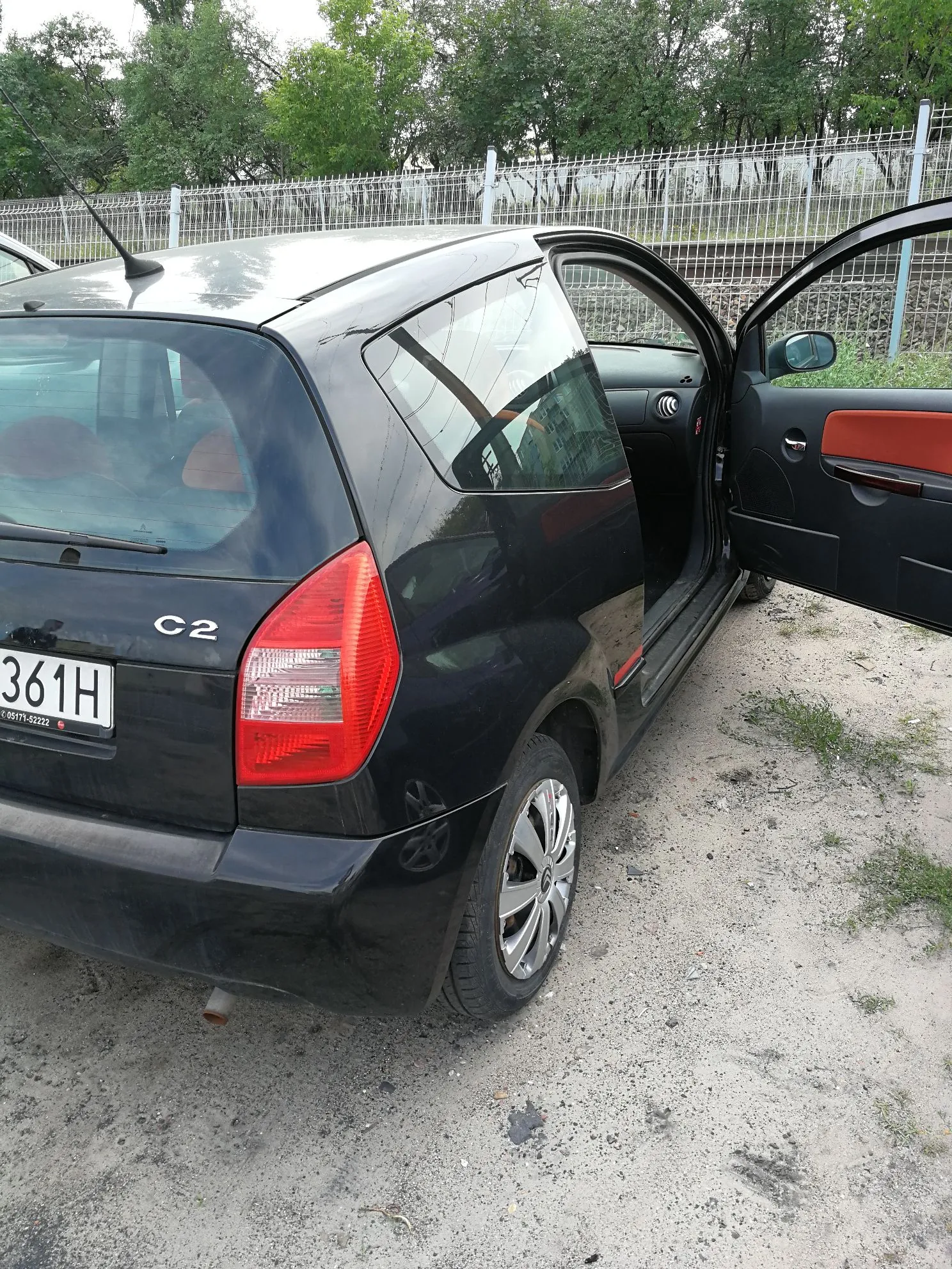 Czarny samochód marki Citroën model C2 zaparkowany na piaszczystym podłożu. Widoczny jest tył i prawy bok auta z otwartymi tylnymi drzwiami, które mają pomarańczowe wykończenie wewnątrz. Samochód posiada tablicę rejestracyjną z numerem 361H. W tle znajduje się metalowy płot oraz zielone drzewa.
