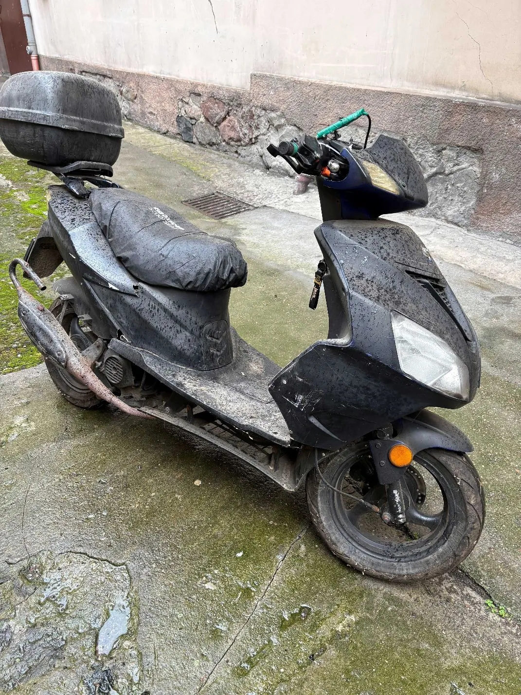 Czarny, zabrudzony skuter zaparkowany na betonowym podłożu obok ściany budynku. Pojazd posiada zamontowany z tyłu czarny kufer. Skuter jest pokryty warstwą kurzu i błota, a rura wydechowa wykazuje oznaki rdzy.