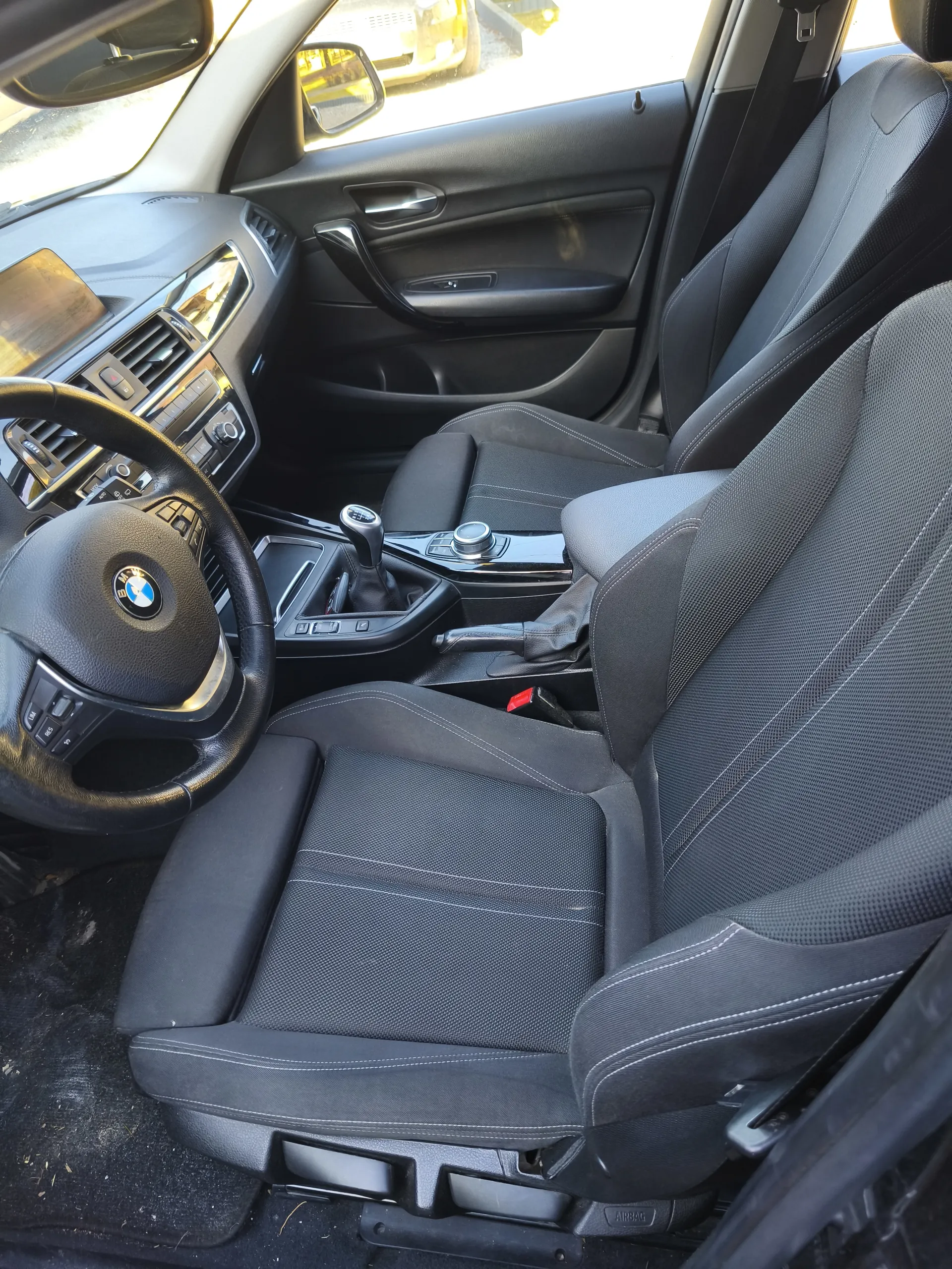 Wnętrze samochodu marki BMW z widocznym kierownicą z logo, czarnymi siedzeniami z białą przeszyciem, centralną konsolą z dźwignią zmiany biegów, deska rozdzielcza z przyciskami i ekranem, drzwi z uchwytem i sterownikami. Widoczne są również lusterka boczne i fragment zewnętrzny z innym pojazdem w tle.