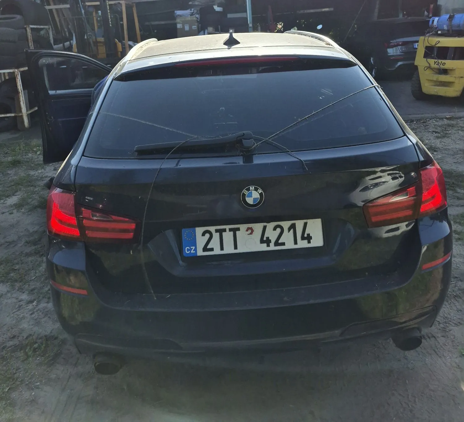 Tylny widok czarnego samochodu marki BMW. Pojazd ma czeską tablicę rejestracyjną o numerze 2TT 4214. Na tylnej szybie widoczne są zarysowania w kształcie litery X. Lewe tylne drzwi są otwarte. W tle po prawej stronie widoczny jest żółty wózek widłowy marki Yale.