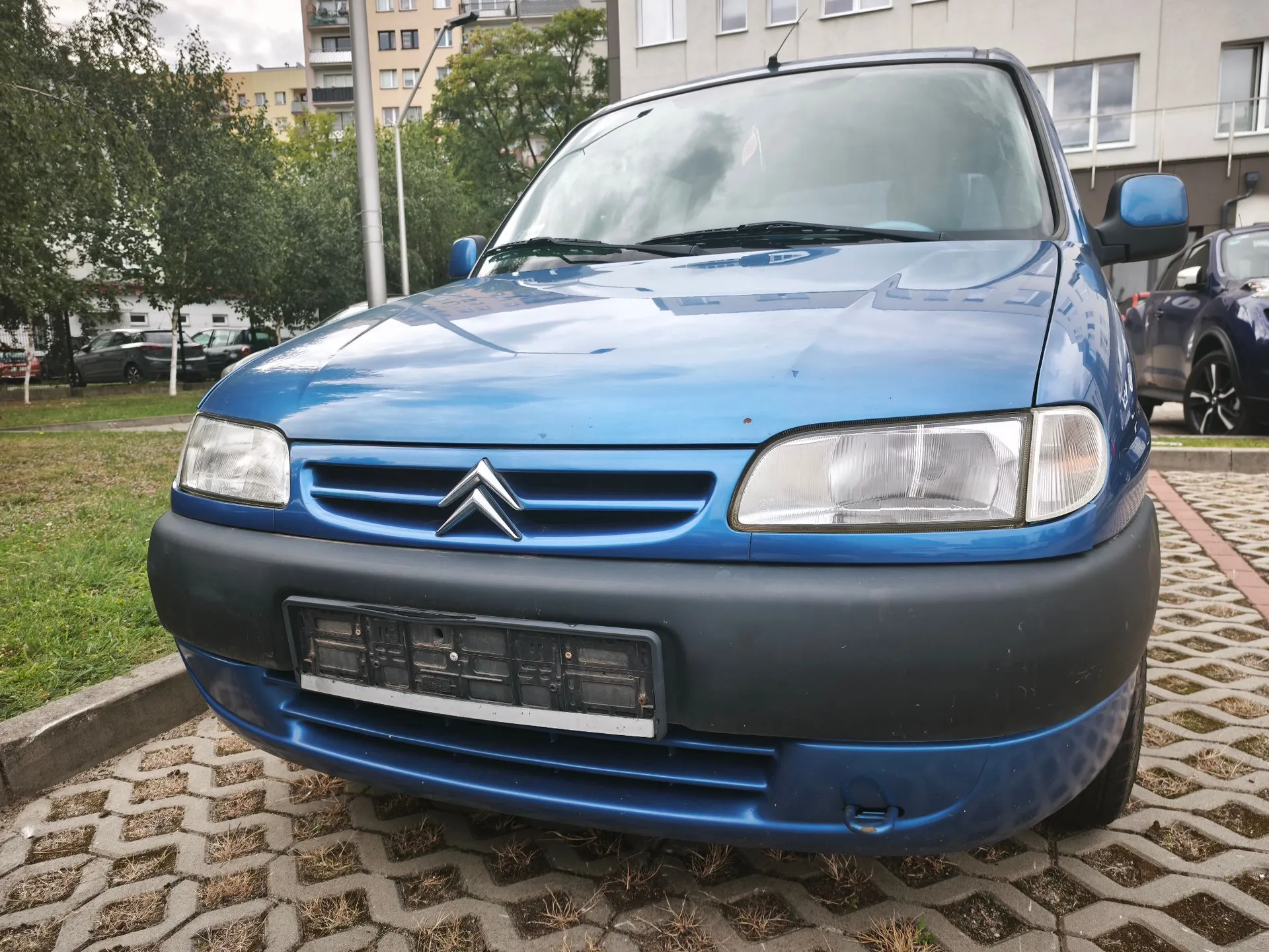 Niebieski samochód marki Citroën zaparkowany na placu z kostki brukowej. Widoczny jest przód pojazdu z charakterystycznym logo w kształcie dwóch kątów (chevronów) na grillu, reflektorami oraz czarnym zderzakiem bez tablicy rejestracyjnej. W tle widać zielone drzewa oraz jasny budynek mieszkalny.