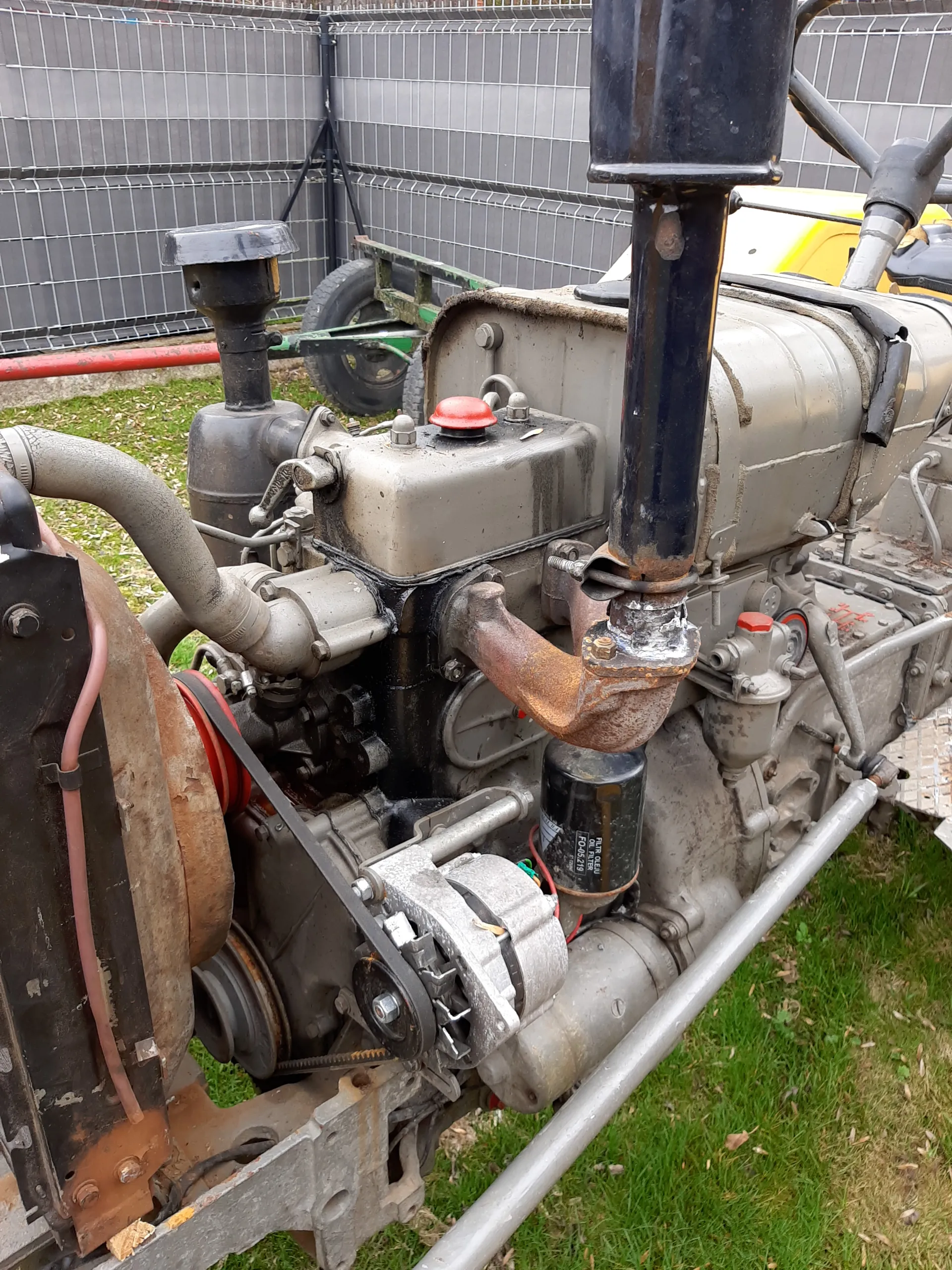 Zbliżenie na silnik z metalowymi częściami, rurami, czarnym cylindrycznym elementem i czerwonymi przyciskami. Widoczny filtr oleju z napisem 'FILTR OLEJU OIL FILTER FO-05219'. W tle trawa i metalowa siatka.
