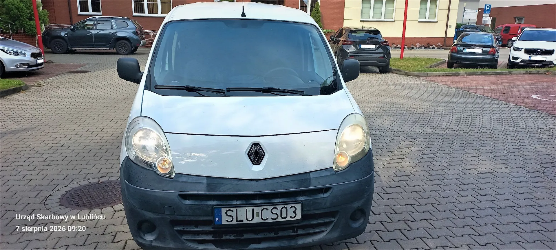 Zdjęcie przedstawiające biały samochód dostawczy marki Renault widziany od przodu, zaparkowany na kostce brukowej. Pojazd ma czarny zderzak i tablicę rejestracyjną o numerze SLU CS03. W tle widoczne są inne zaparkowane samochody oraz budynek. W lewym dolnym rogu znajduje się napis: 'Urząd Skarbowy w Lublińcu 7 sierpnia 2026 09:20'.