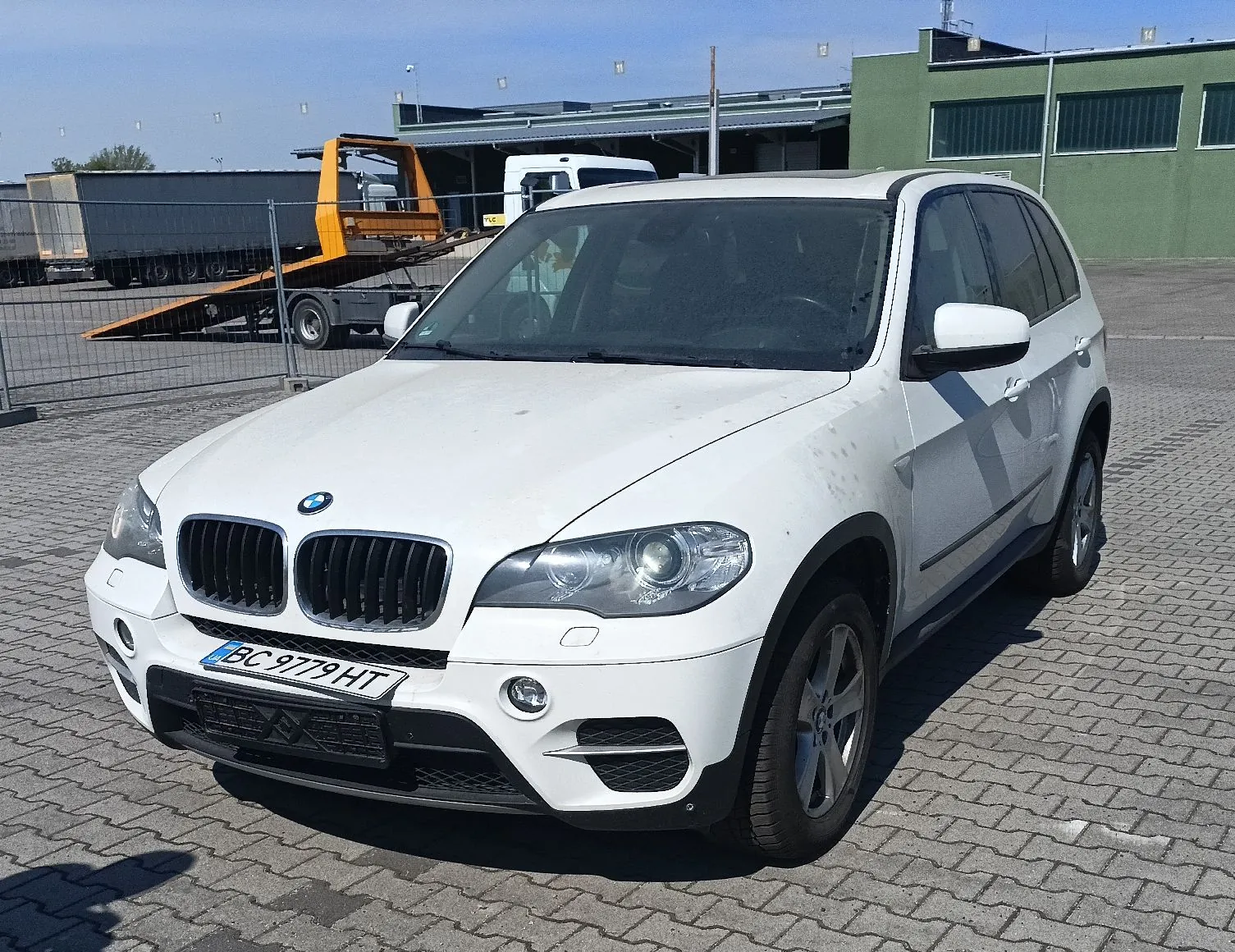 Biały samochód marki BMW X5 zaparkowany na placu wyłożonym kostką brukową. Pojazd ma tablicę rejestracyjną o numerze BC 9779 HT. W tle widoczny jest żółty samochód laweta oraz zielony budynek przemysłowy pod jasnym, niebieskim niebem.