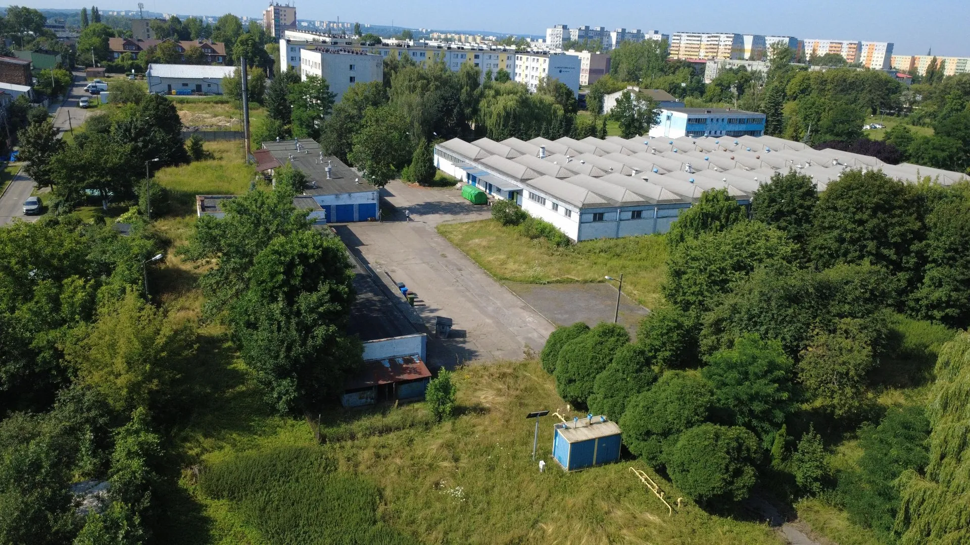 Widok z lotu ptaka na teren przemysłowo-biurowy otoczony gęstą roślinnością i drzewami. W centrum znajduje się duży, biały budynek magazynowy z charakterystycznym ząbkowanym dachem. Obok niego znajduje się betonowy plac manewrowy oraz mniejsze budynki gospodarcze. W tle widoczne są wielopiętrowe bloki mieszkalne i zabudowania miejskie pod jasnym niebem.