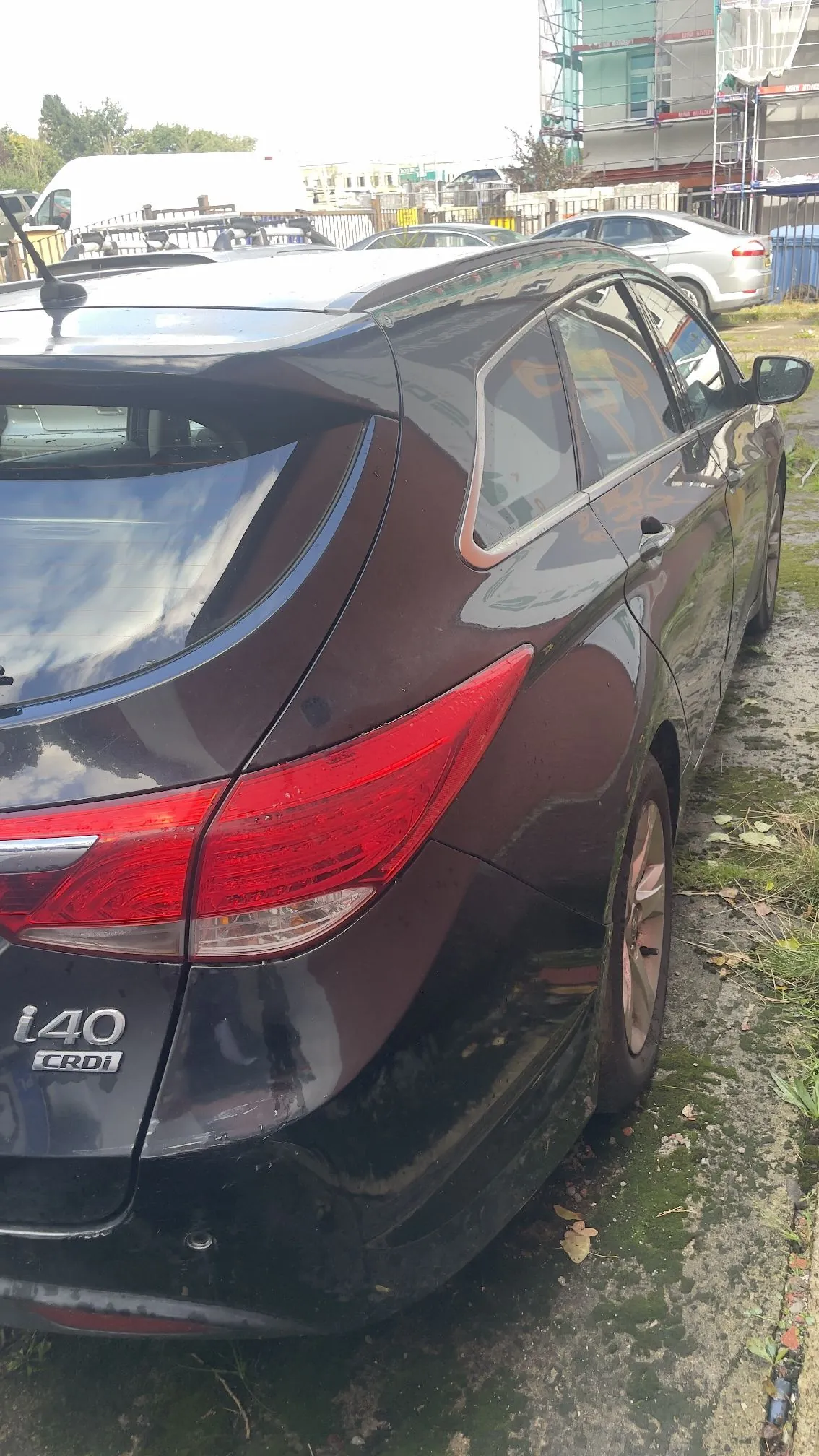 Tylna i boczna część czarnego samochodu marki Hyundai i40 CRDi. Pojazd jest zaparkowany na utwardzonym podłożu z mchem. W tle widoczne są inne samochody oraz budynek w trakcie remontu z rusztowaniami.