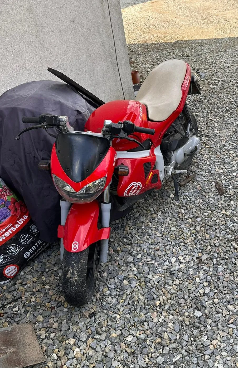 Czerwony skuter z beżowym siedzeniem i białymi logo na boku, zaparkowany na żwirowym podłożu. Obok skutera znajduje się czarna pokrowiec oraz worek z napisem 'ziemia uniwersalna' i 'FERTIL'. W tle betonowa ściana i żwirowa powierzchnia.