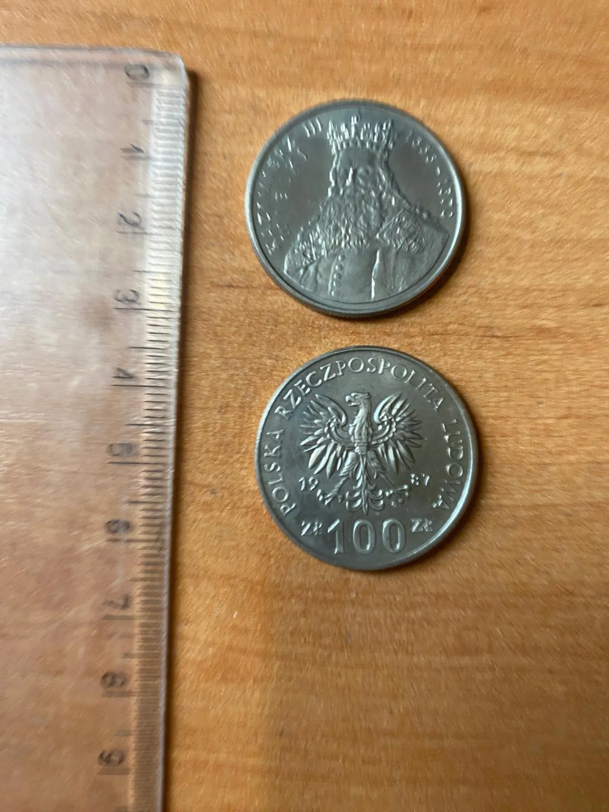 Zdjęcie przedstawiające dwie strony srebrzystej monety leżącej na drewnianym blacie obok metalowej linijki. Górna moneta przedstawia profil postaci w koronie. Dolna moneta zawiera napis 'POLSKA RZECZPOSPOLITA LUDOWA', rok '1975' oraz nominał '100 zł' z wizerunkiem orła w centrum.